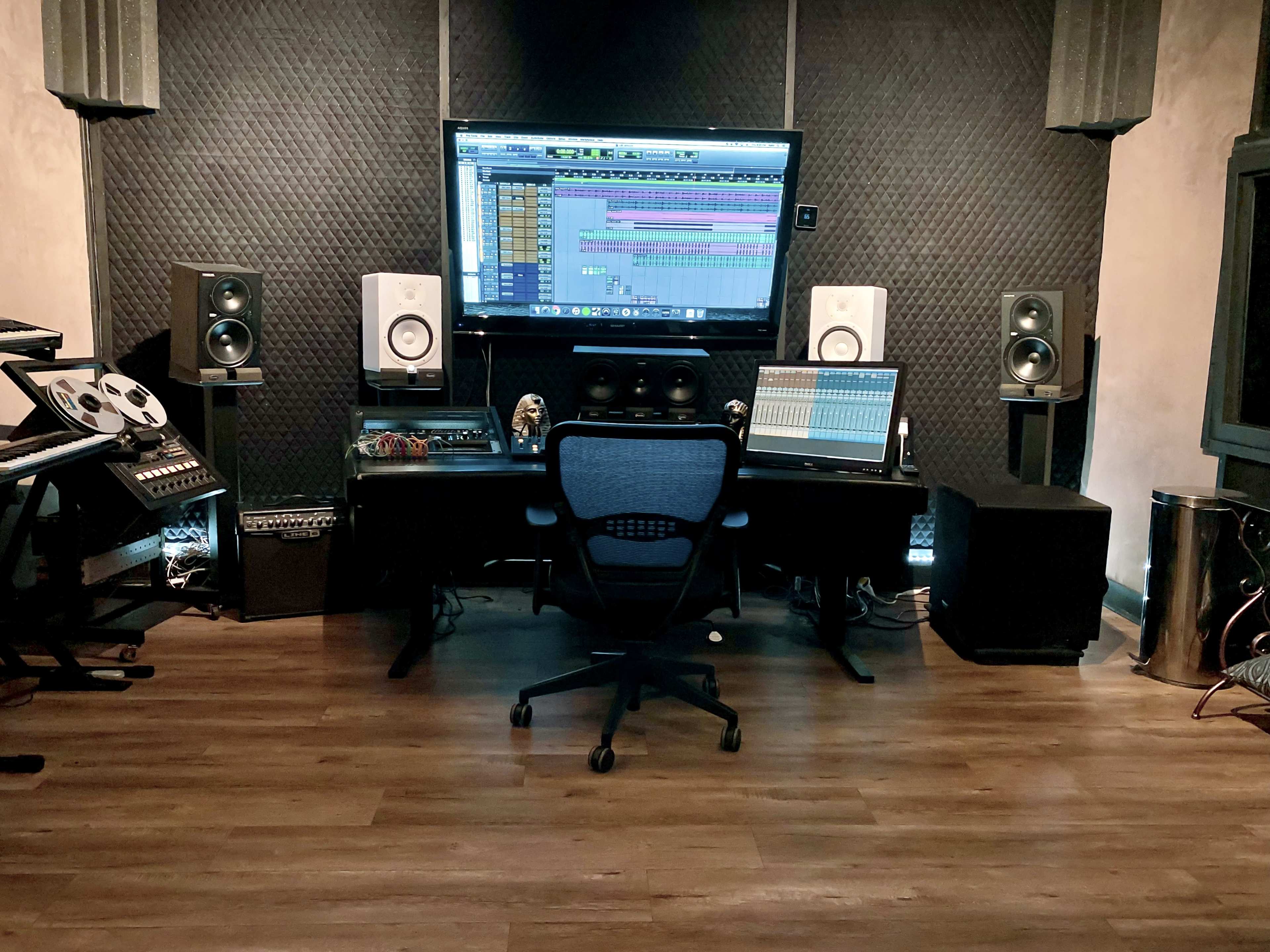 A music studio featuring a computer monitor displaying audio software, a digital audio workstation, studio speakers, and various musical equipment arranged around a central swivel chair.