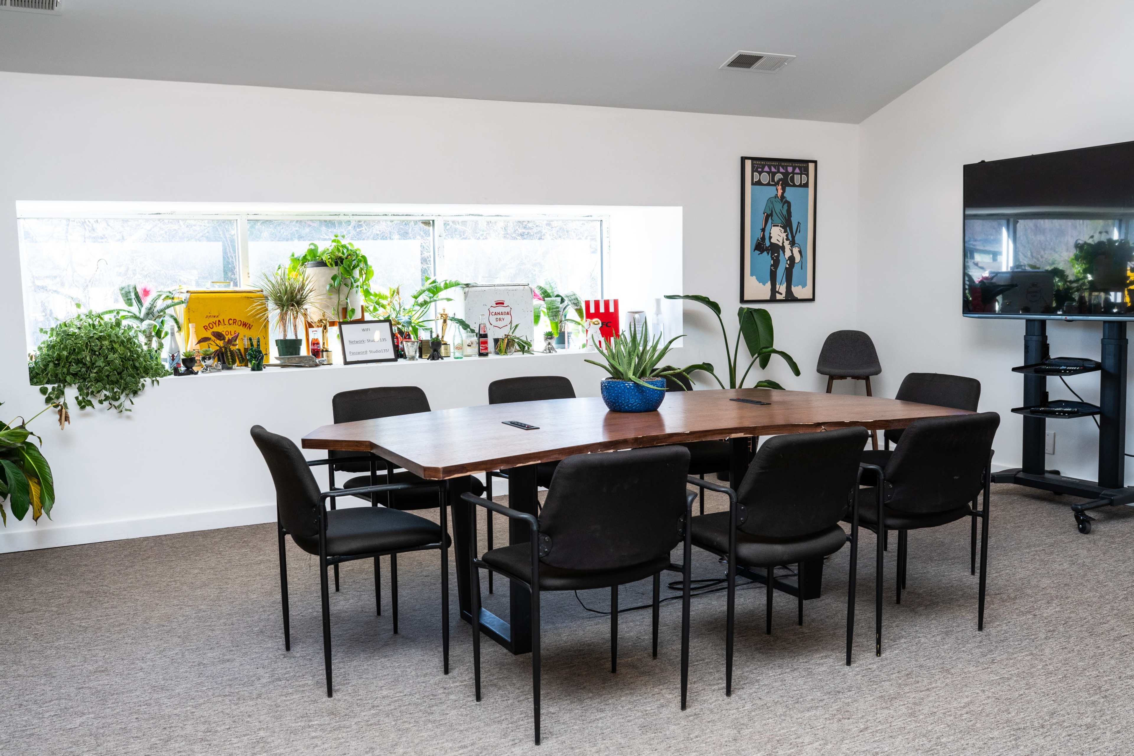 A long wooden table surrounded by black chairs is positioned in a well-lit meeting room with potted plants and decorative items on the windowsill.