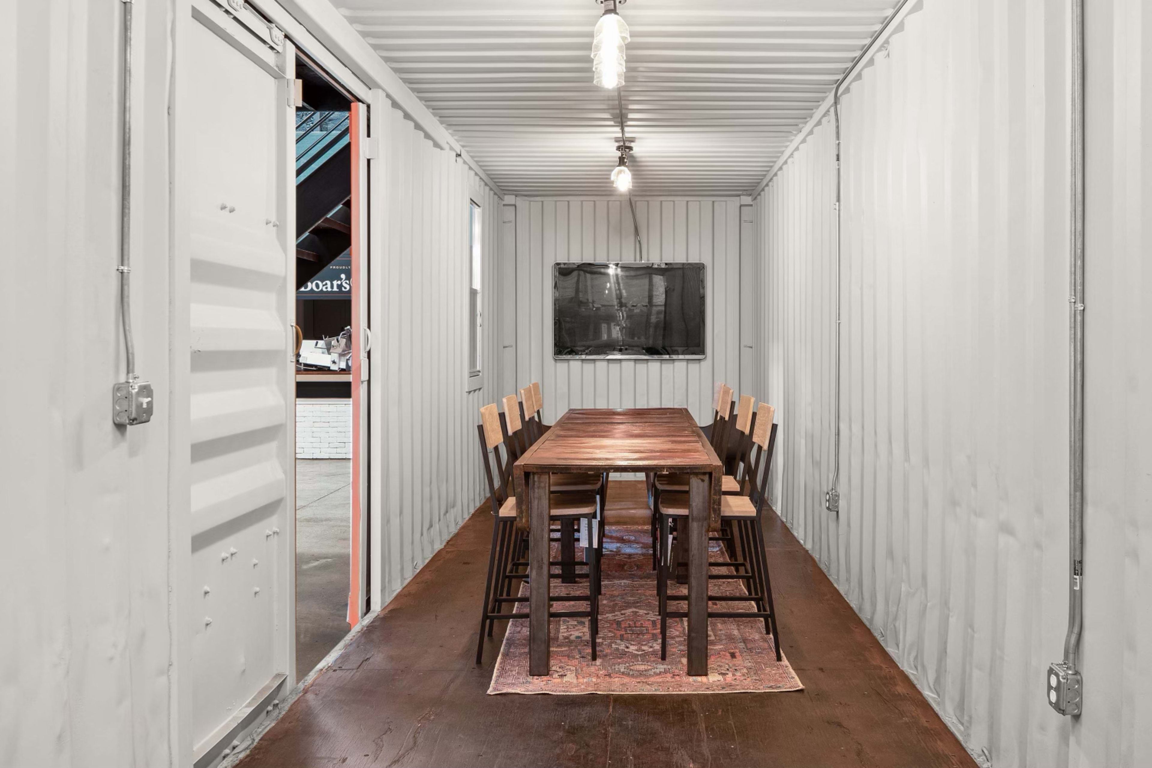 The image shows a minimalist dining area inside a shipping container, featuring a long wooden table with eight chairs and a television mounted on the wall.