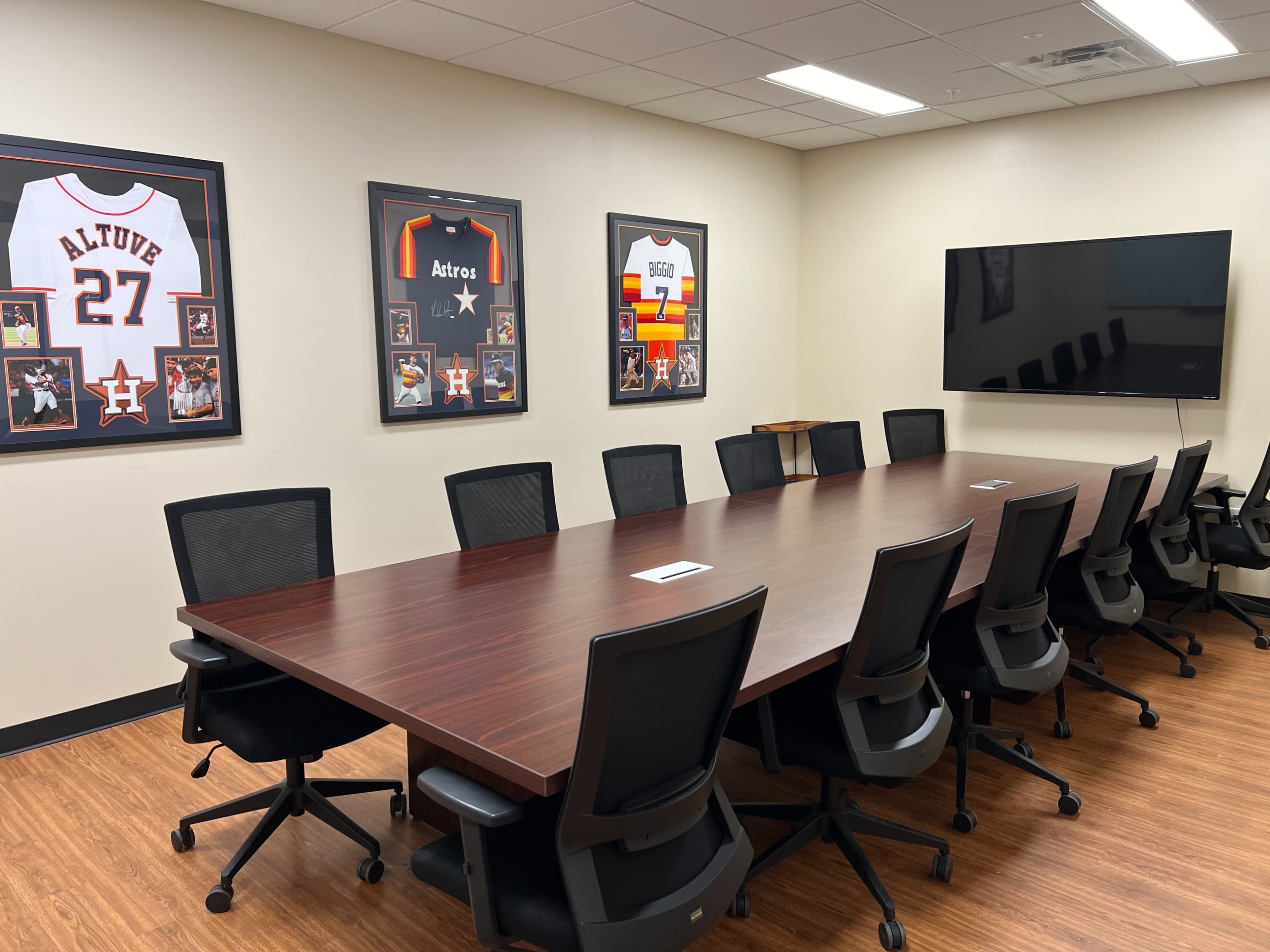 A conference room with a long wooden table surrounded by black office chairs, adorned with framed jerseys and a large television on the wall.