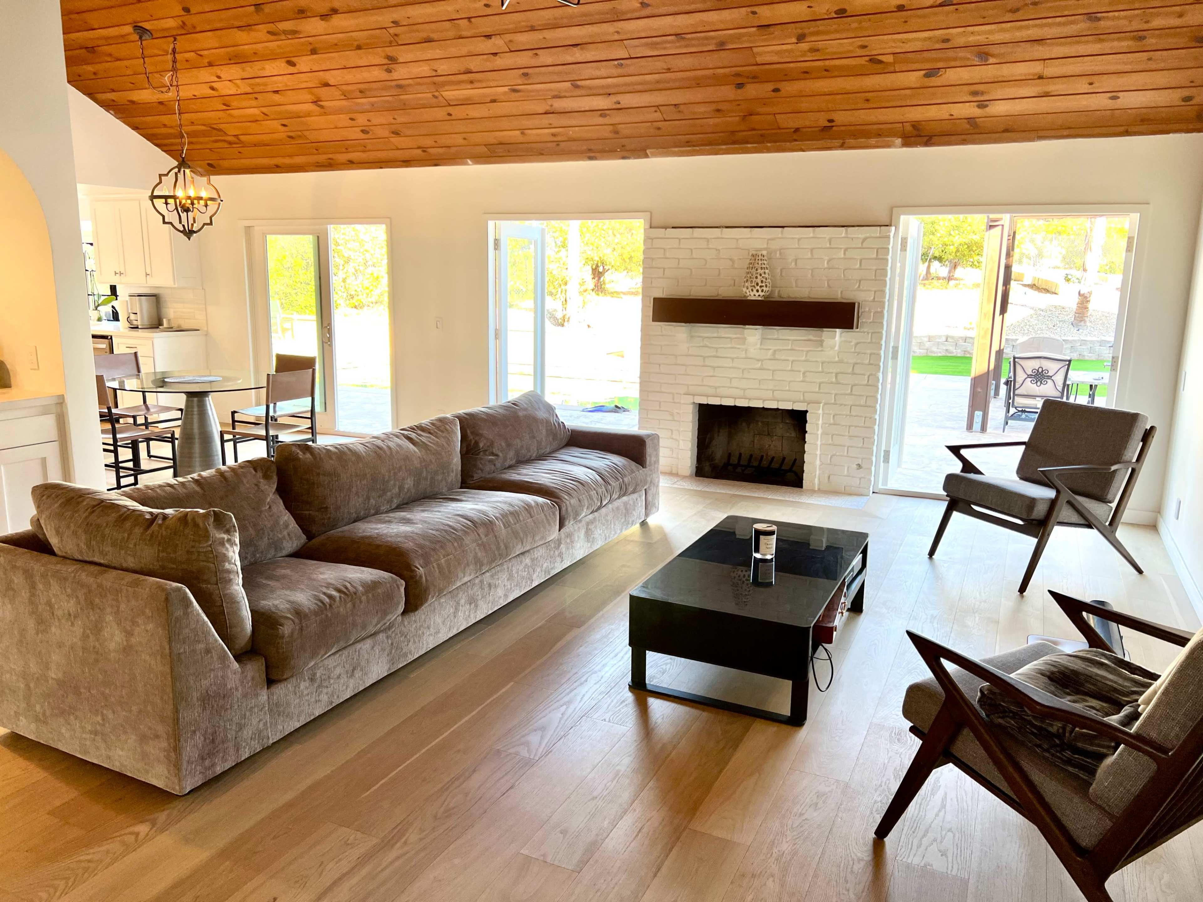 The living room features a large gray sofa, a glass coffee table, and two armchairs, with a white brick fireplace and large windows overlooking an outdoor space.