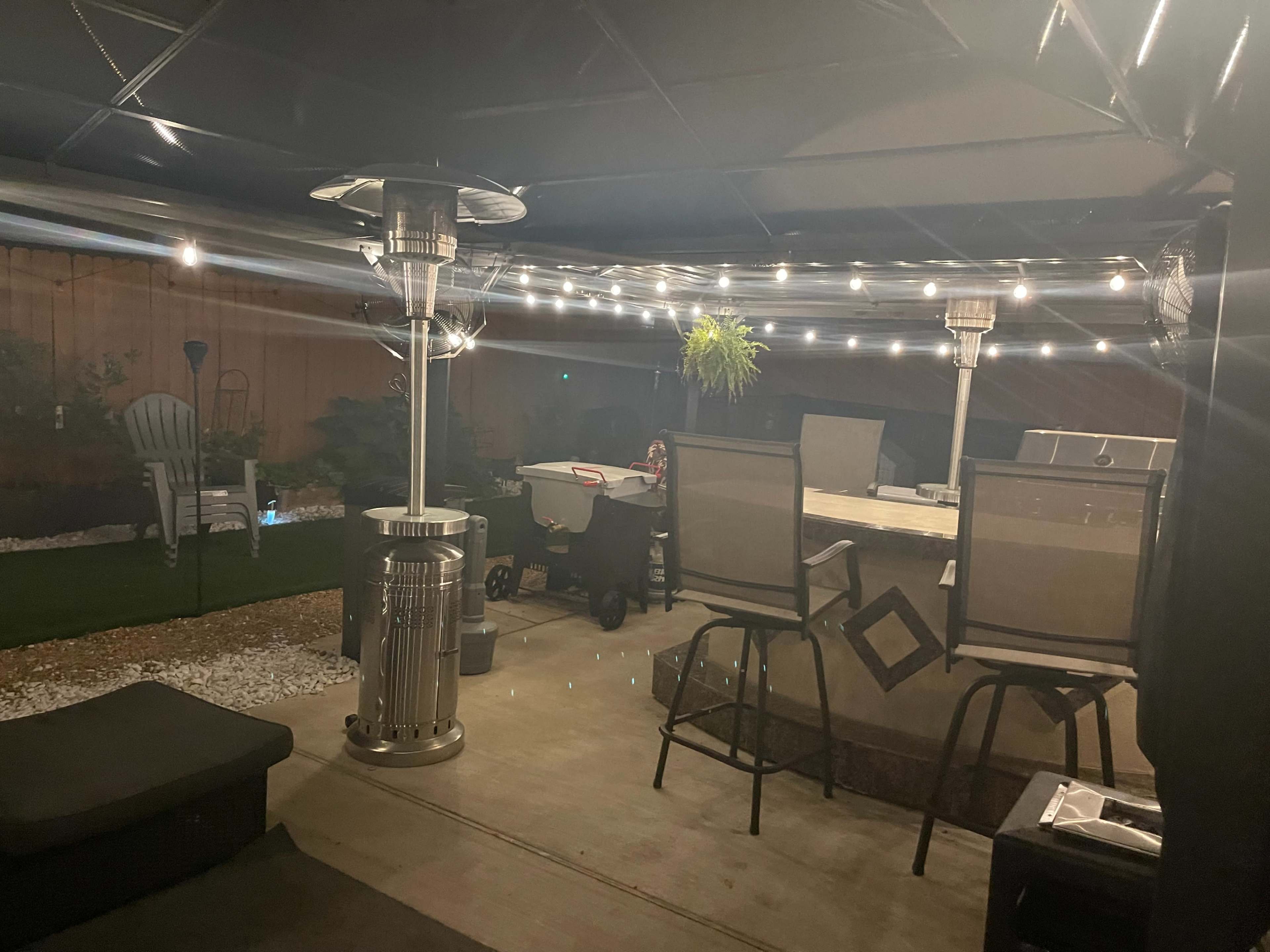 The image shows a patio area with outdoor heaters, a dining table surrounded by chairs, string lights overhead, and a grassy section with landscaping.