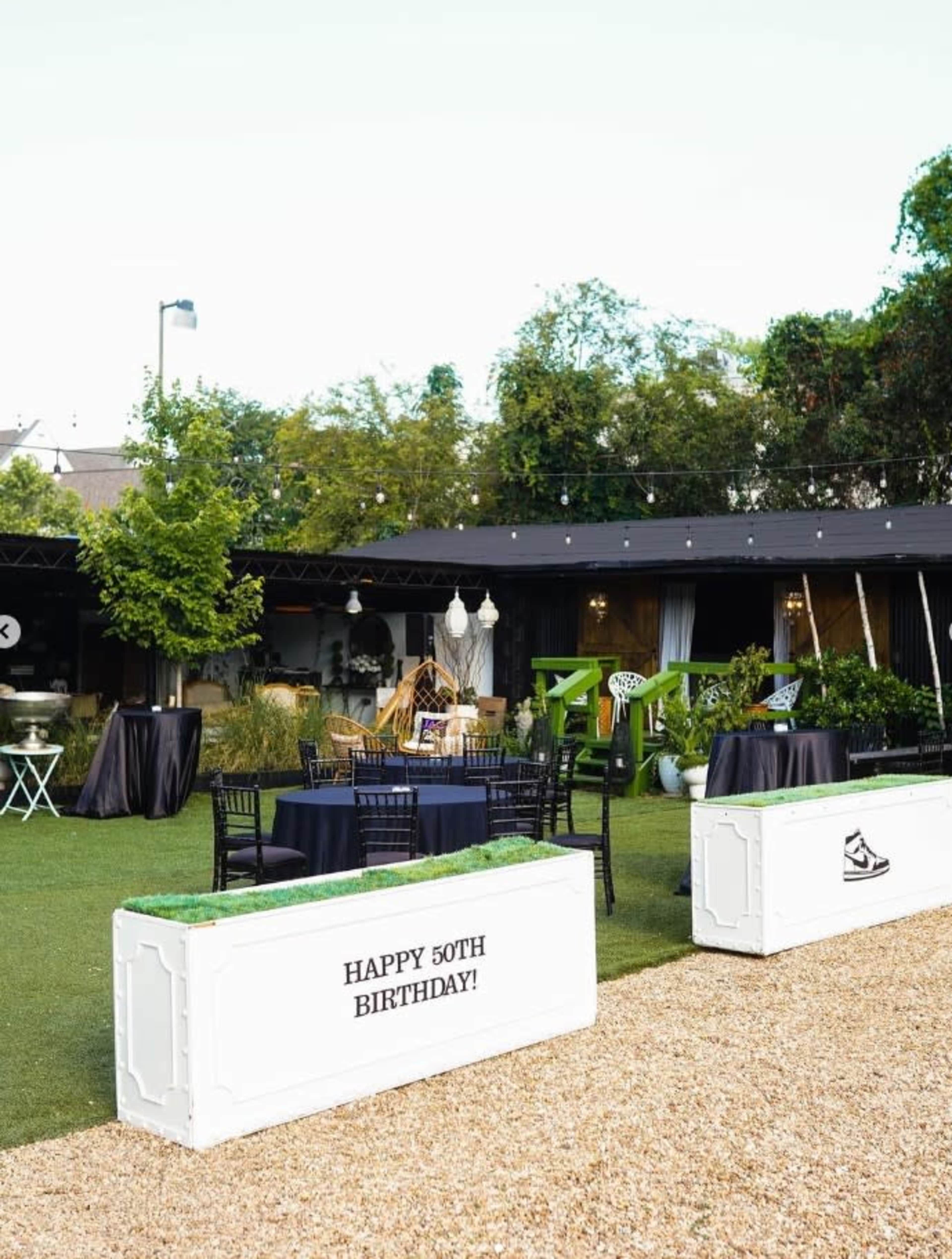 The image shows a garden setting for a 50th birthday celebration, featuring decorated tables and a large banner that reads "Happy 50th Birthday!"