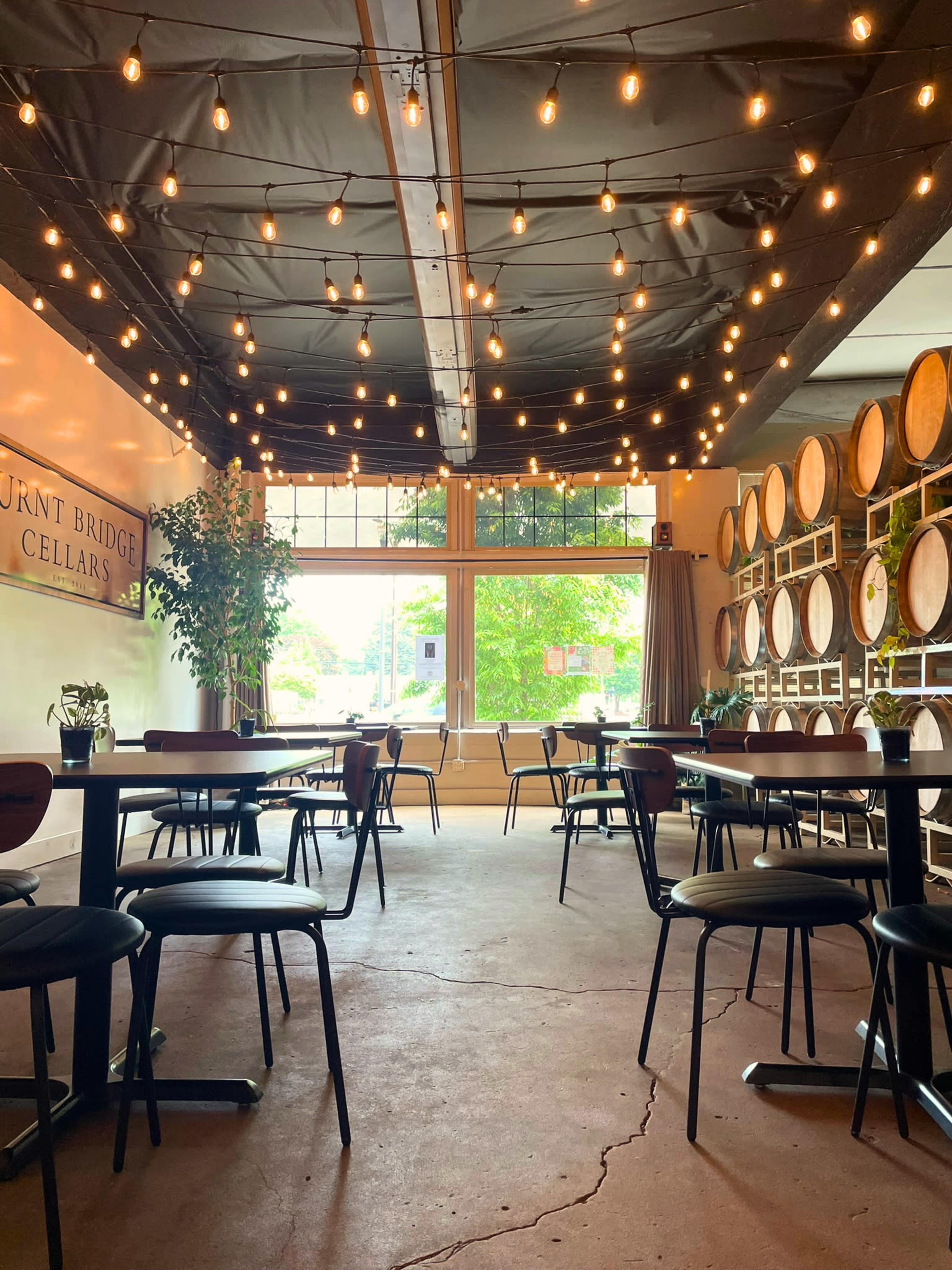 The interior of a wine cellars tasting room features round tables and chairs arranged on a concrete floor, with large windows and decorative string lights above.