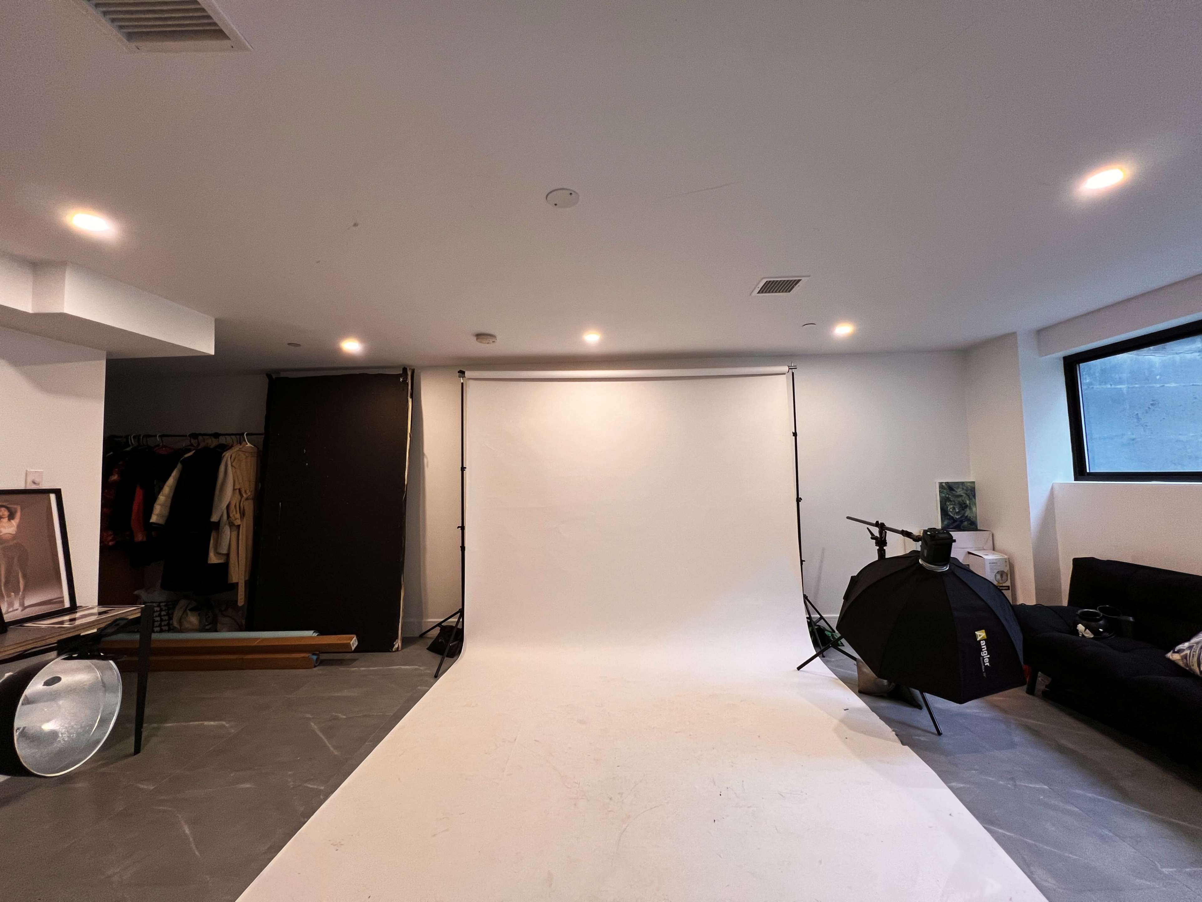 The image shows a photography studio with a blank backdrop, studio lights, a clothing rack, and a seating area.