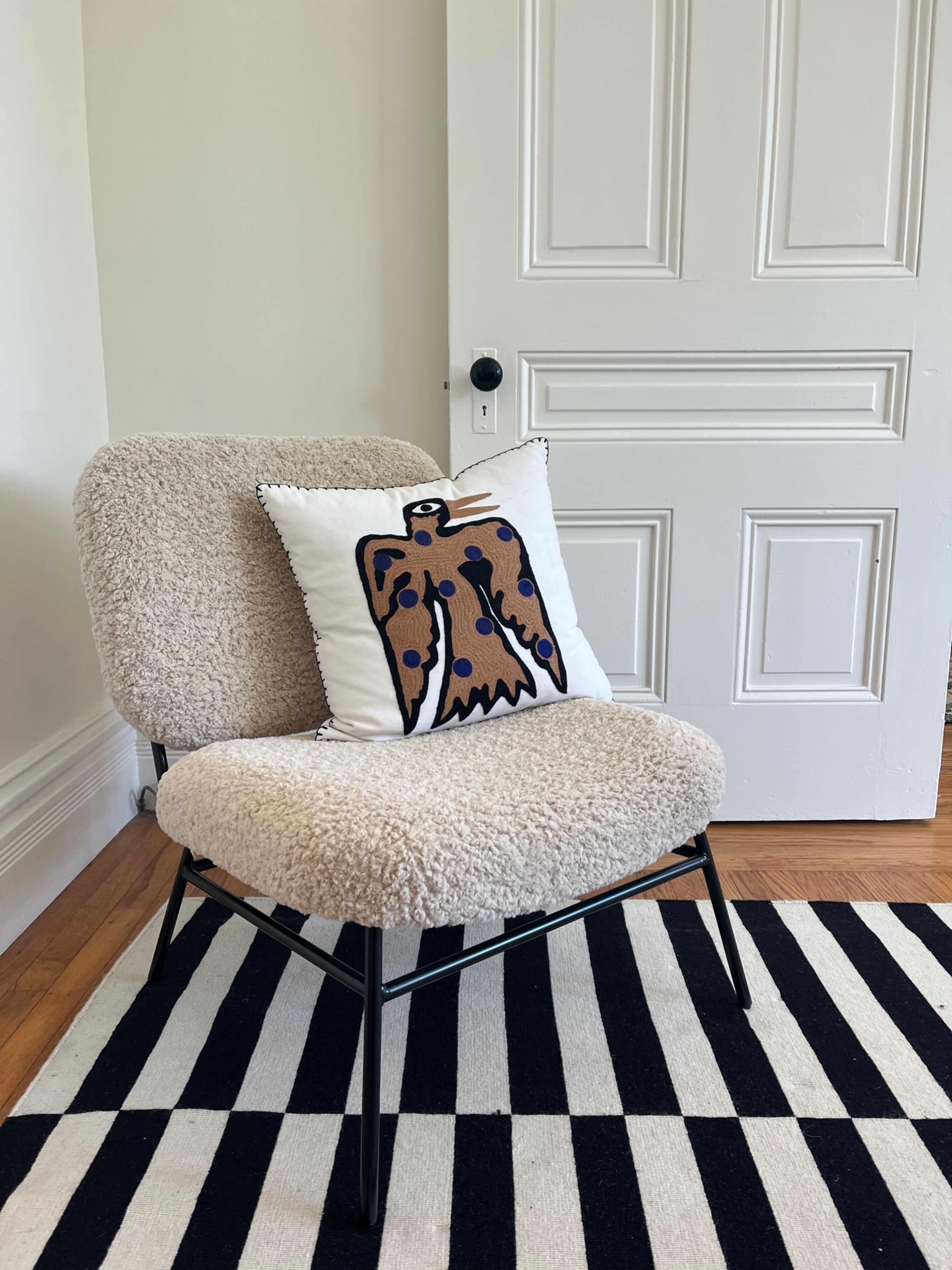 A plush, cream-colored chair with a decorative pillow featuring an abstract design, placed on a black and white striped rug next to a closed white door.