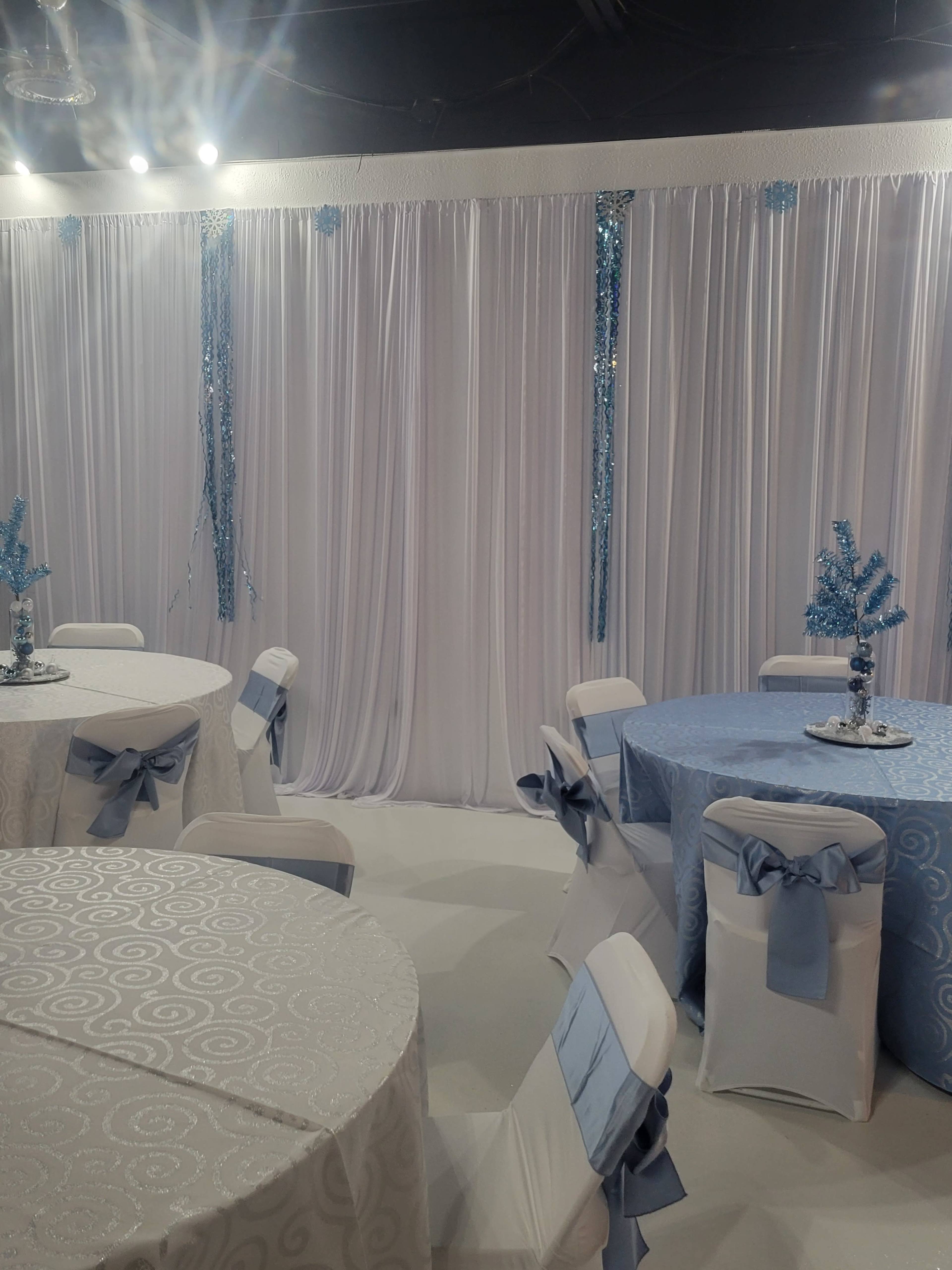 The image shows a decorated event space with white and light blue tablecloths, chairs with light blue sashes, and a backdrop of white drapery accented with blue elements.