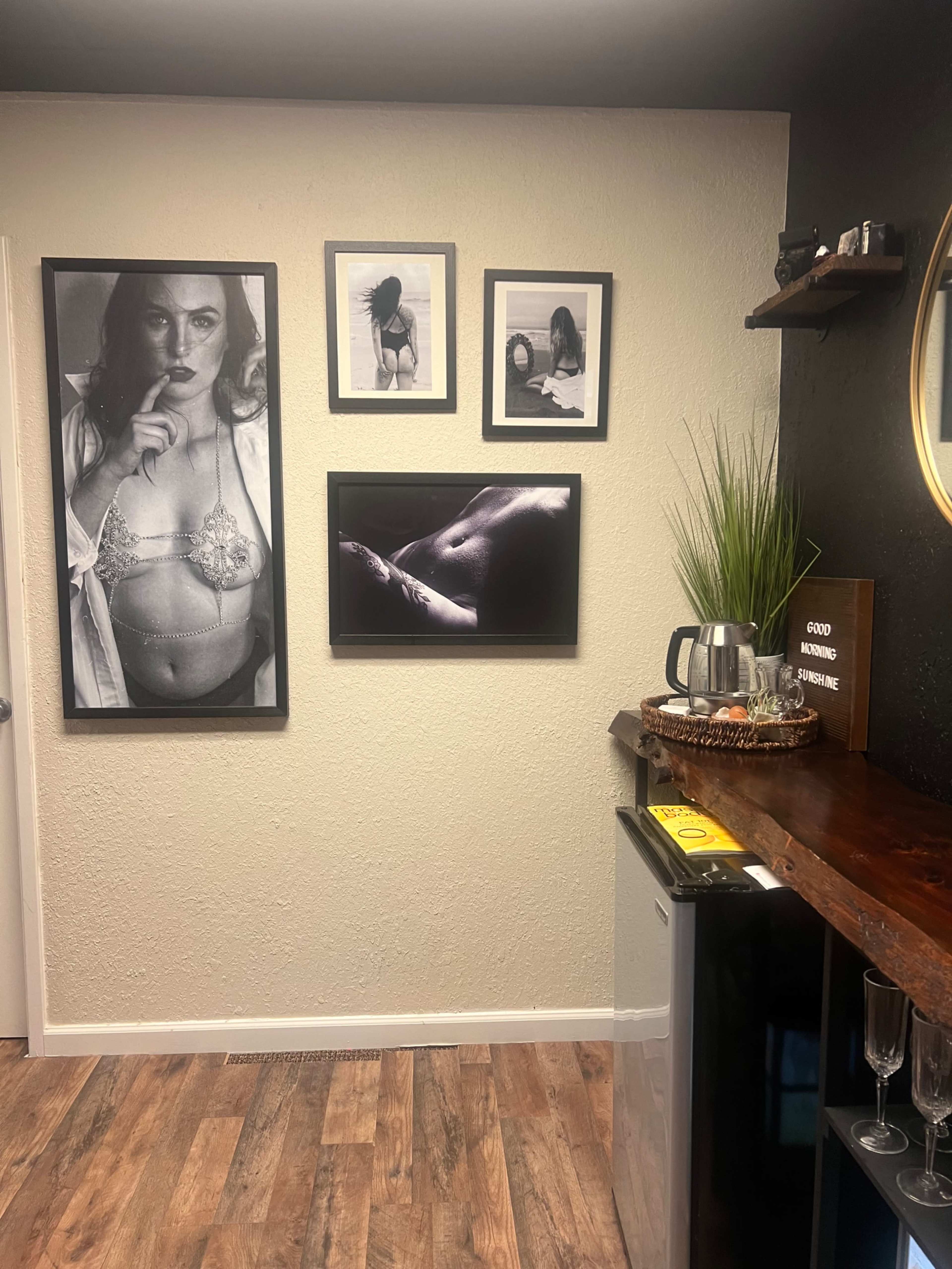 The image shows a wall adorned with framed black-and-white photographs, a round mirror, and various decor items in a small room with wooden flooring.