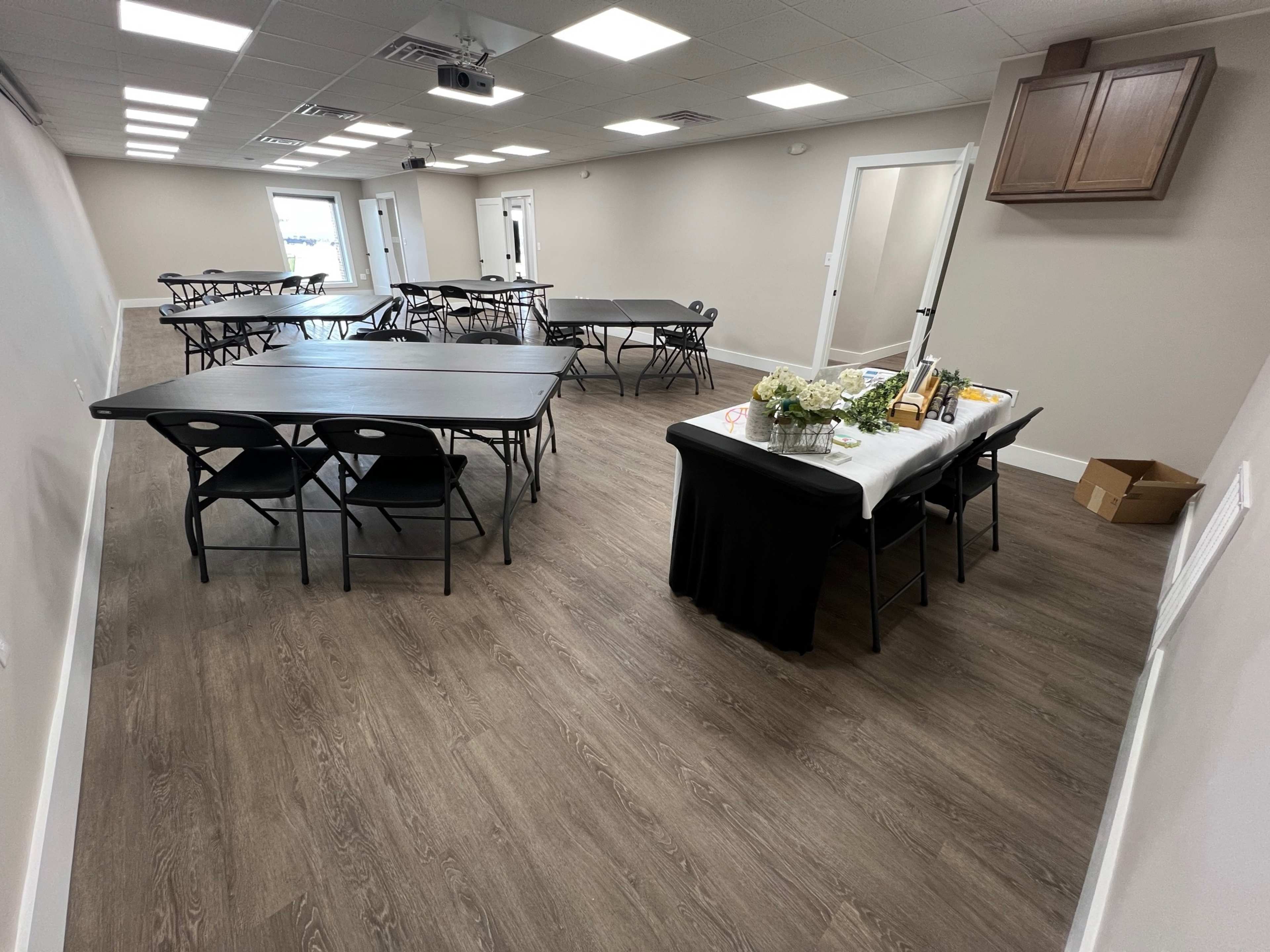 The image shows a spacious, modern room with several black folding tables and chairs arranged for an event, along with a table set up for catering in one corner.