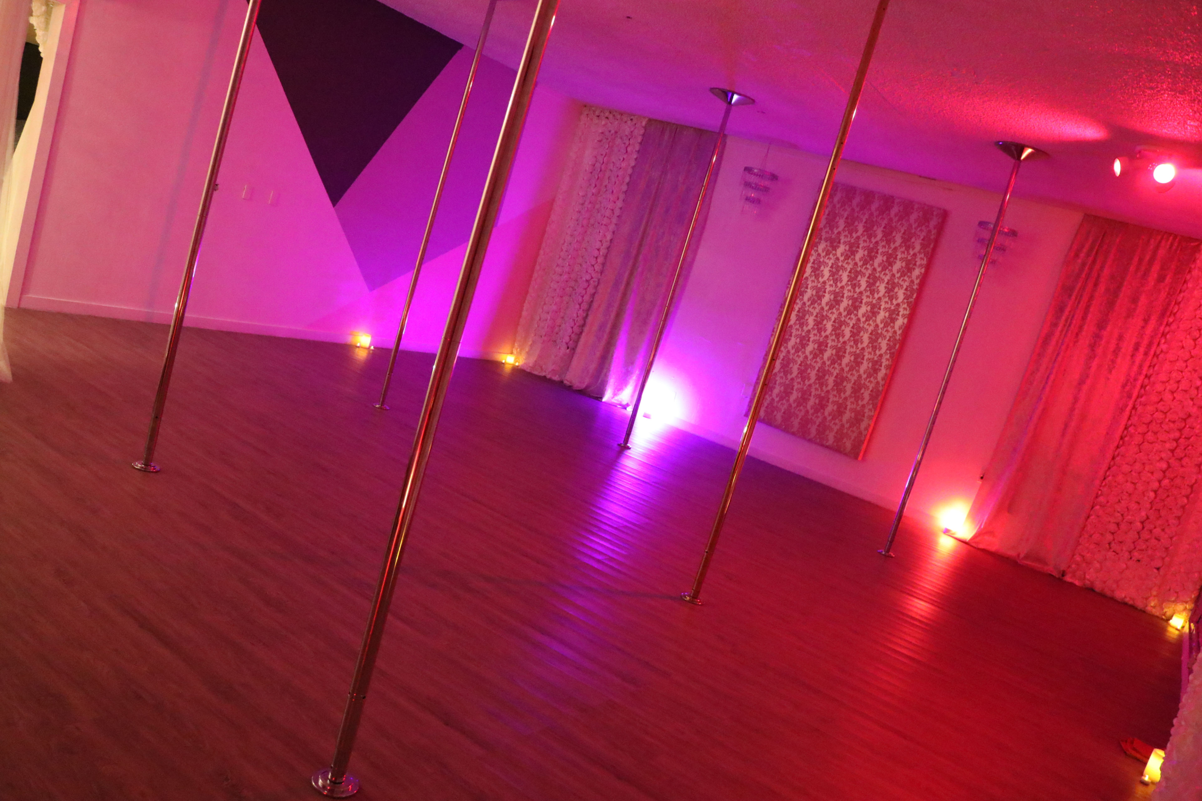 An empty dance studio featuring several vertical poles, with dim pink and purple lighting illuminating the space.