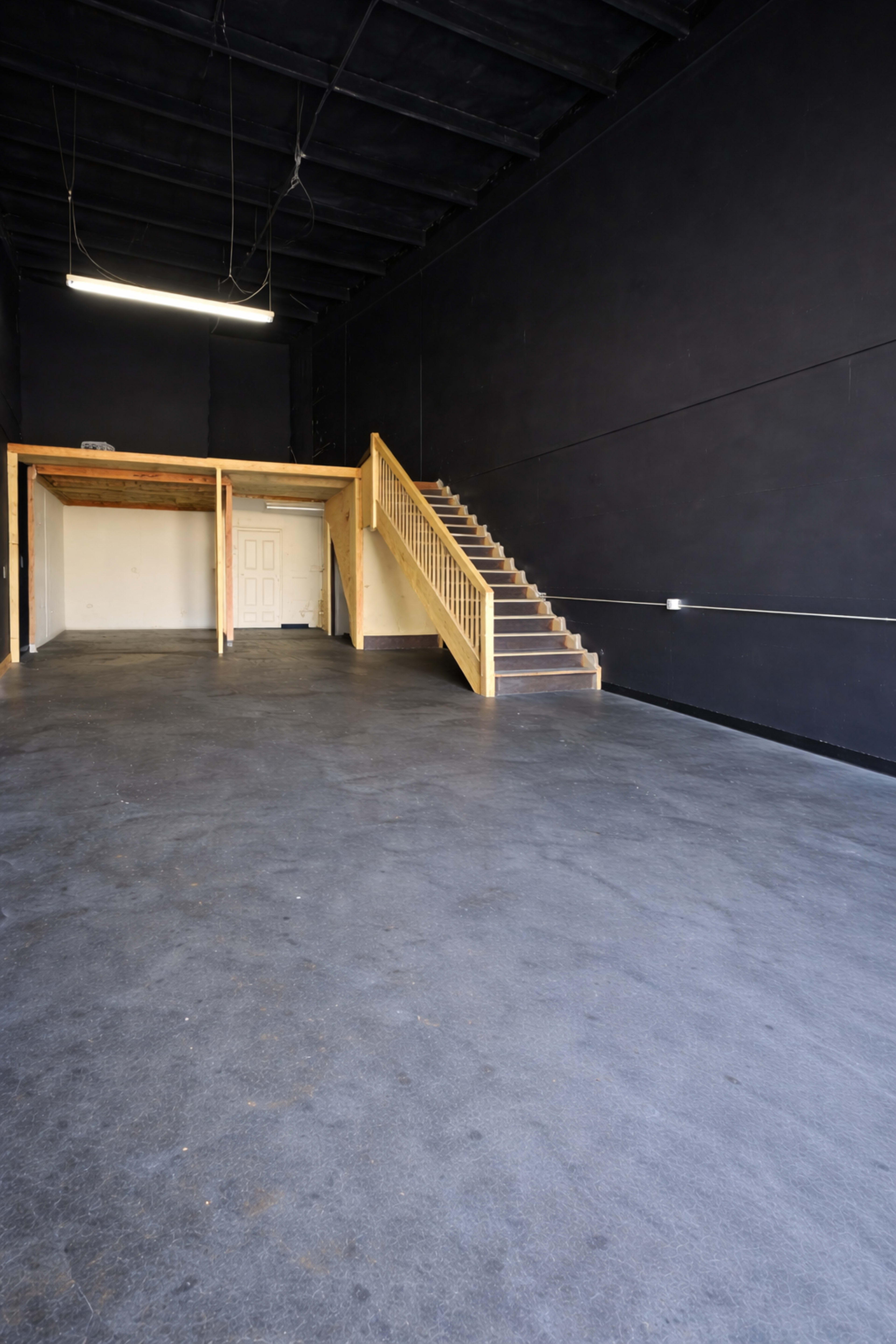 The image shows an empty interior space with dark walls, a wooden staircase on the right, and a small area with a door and platform in the back.