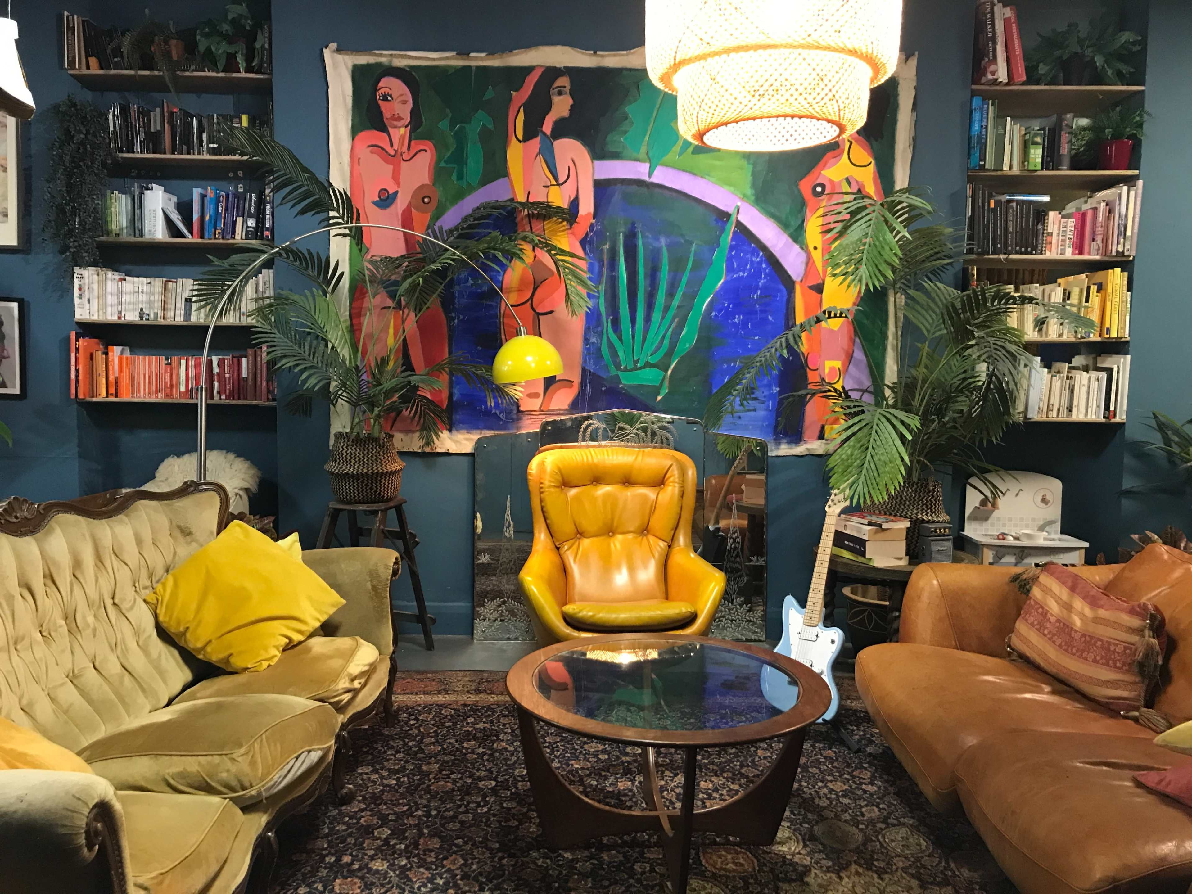 The scene features a cozy living room with a yellow armchair, plants, a round coffee table, and a colorful mural of two figures on the wall.