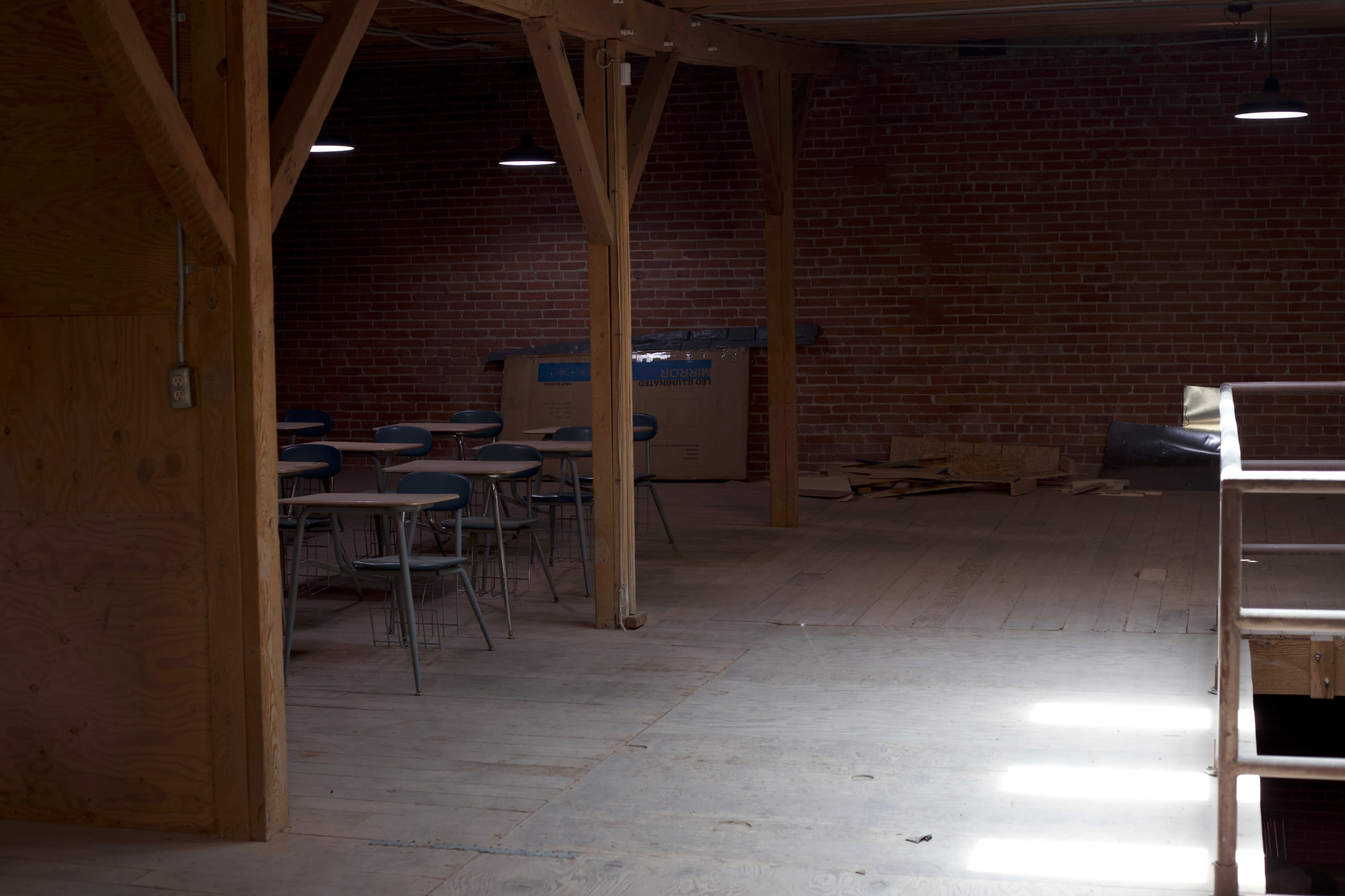 A sparsely furnished room with wooden beams and a few chairs arranged around a table, illuminated by overhead lights.