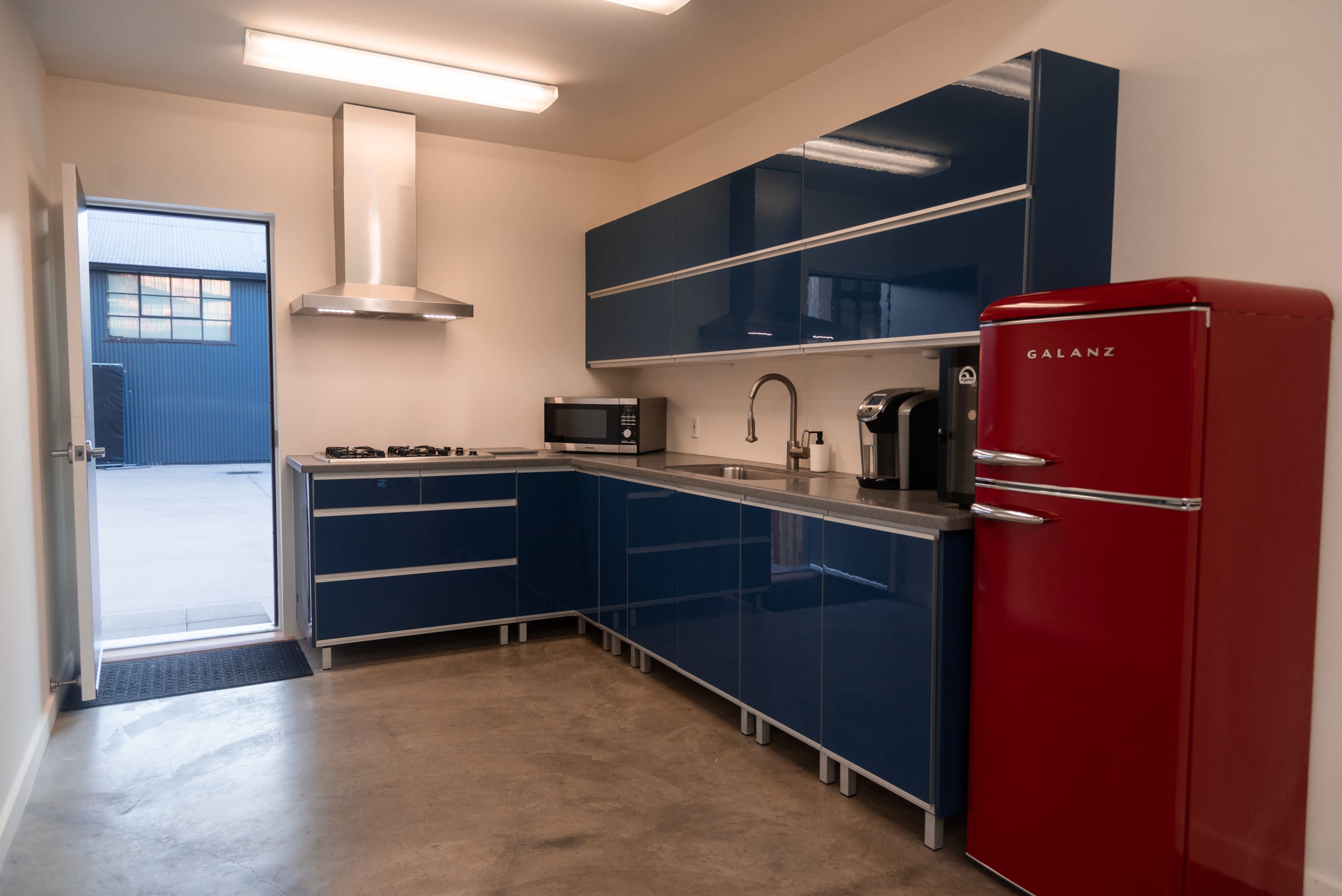 The image shows a modern kitchen featuring glossy blue cabinets, a red retro-style refrigerator, stainless steel appliances, and a concrete floor.