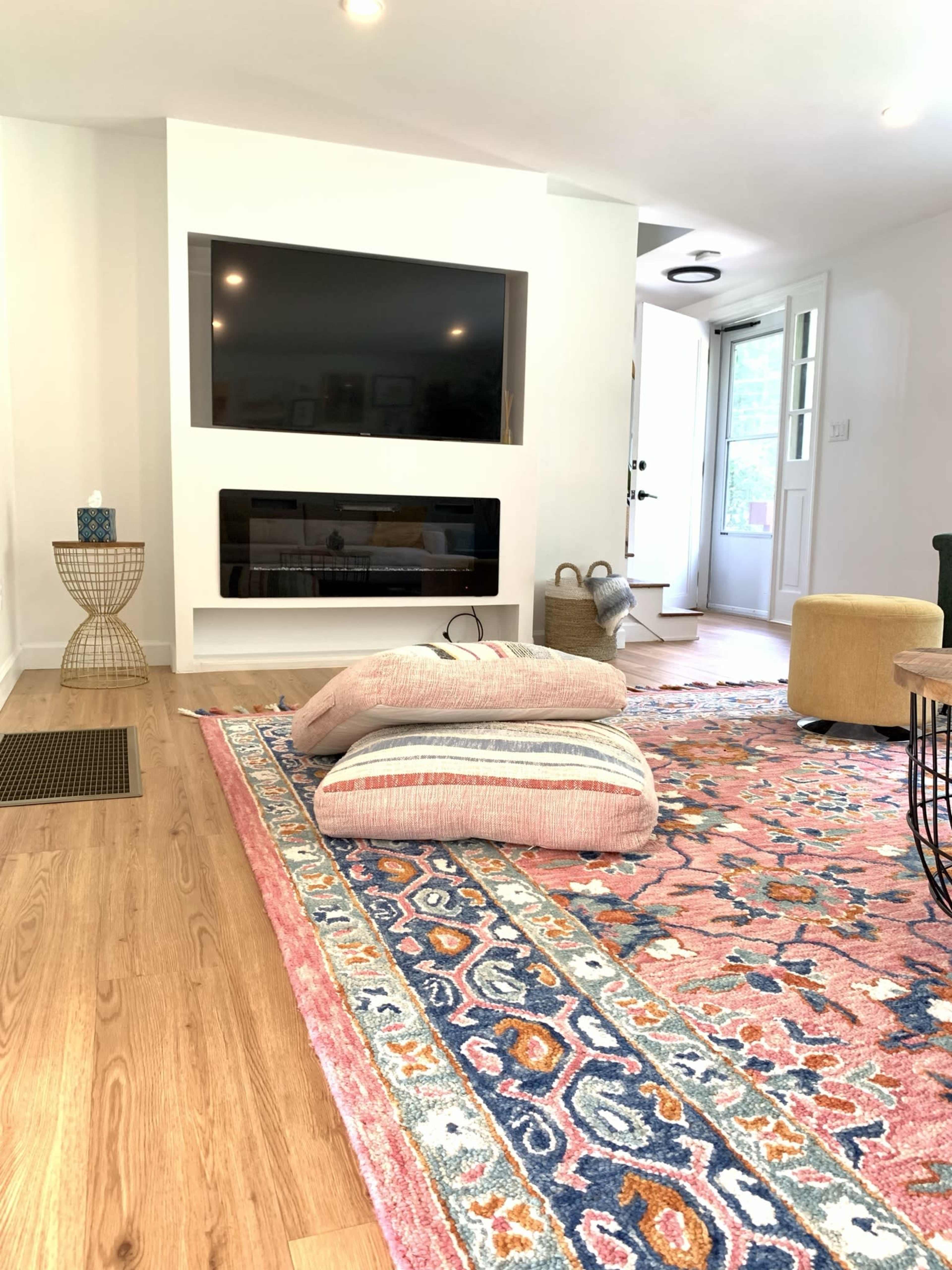 The scene shows a modern living room with a large flat-screen TV mounted on the wall, colorful patterned rugs on a wooden floor, and a few casual cushions scattered on the rug.