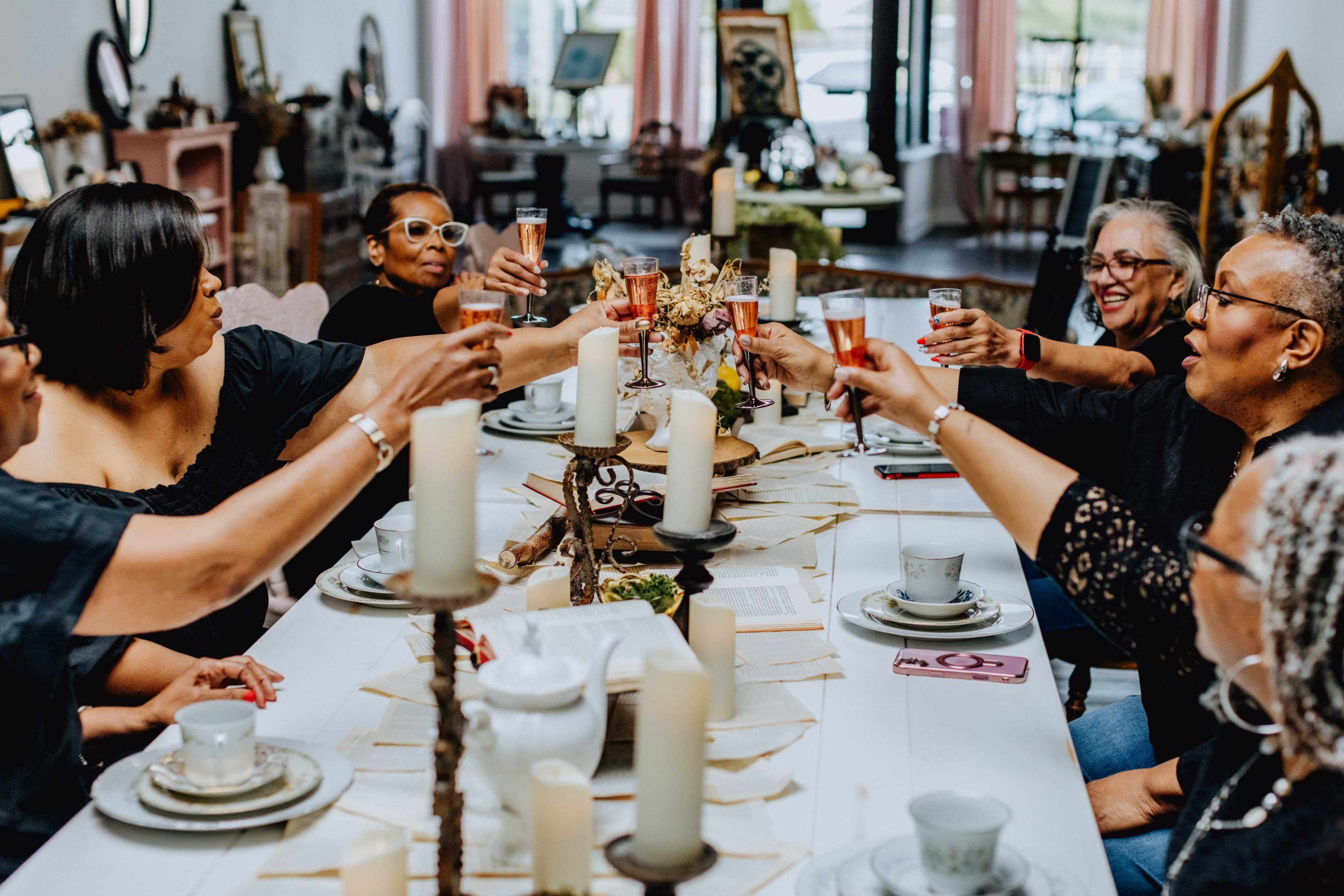 A group of women toast with drinks at a long table set with candles and tableware in a decorated indoor space.