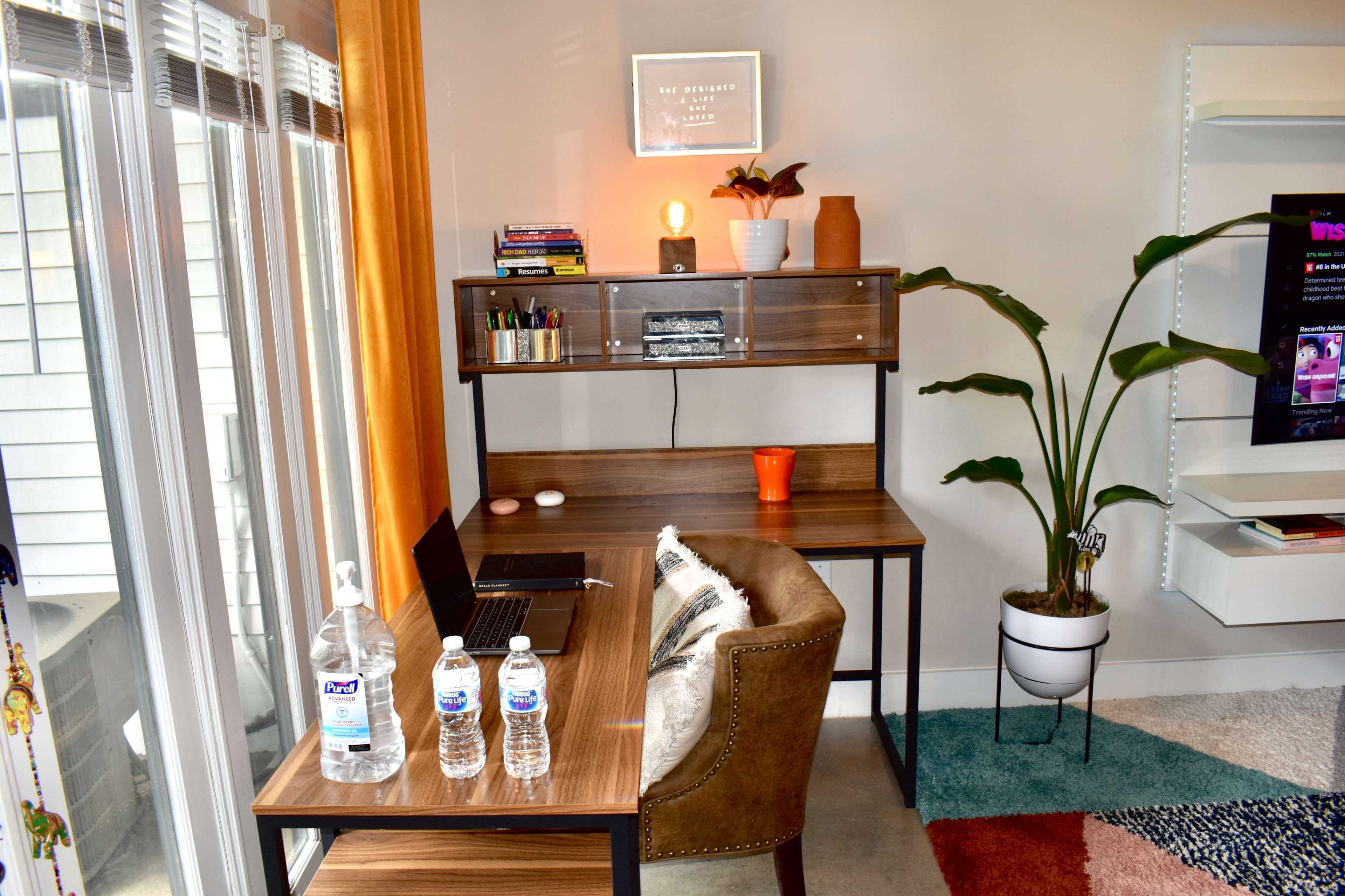 A home office setup featuring a wooden desk with a laptop, three water bottles, and a decorative plant beside it, set against a light-colored wall with large windows.
