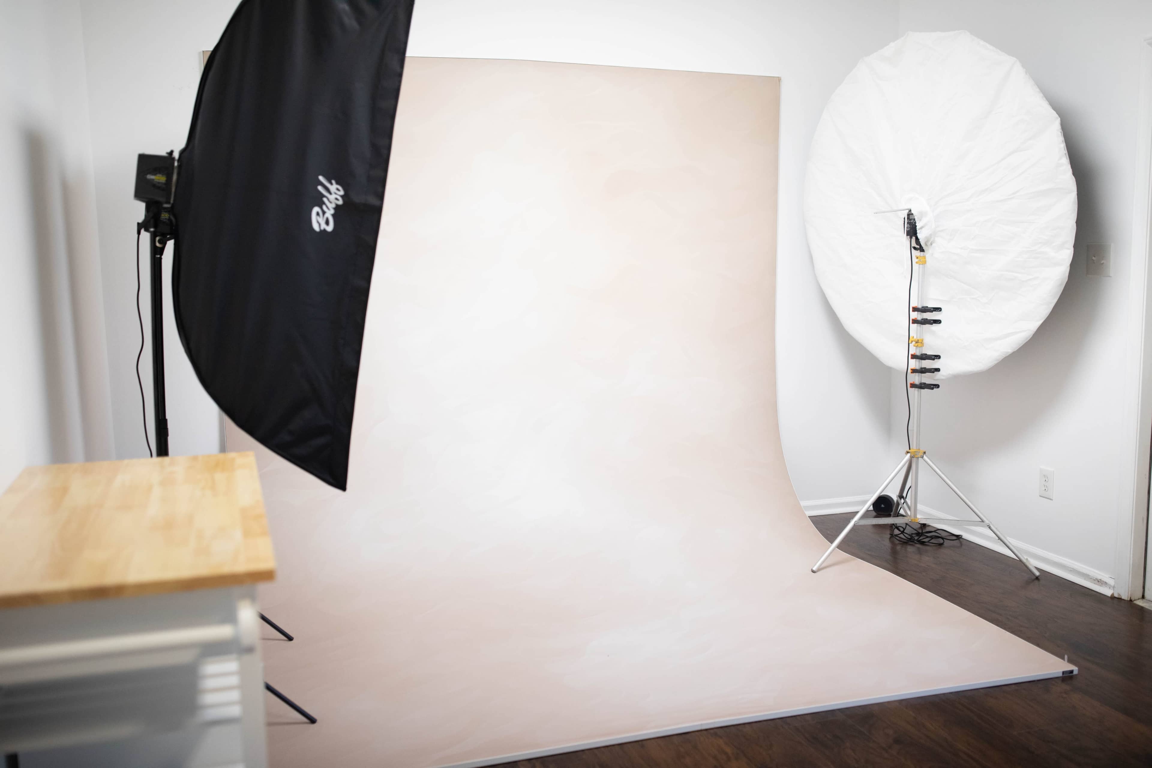 The image shows a photography studio setup featuring a large light modifier to the left, a circular softbox to the right, and a plain backdrop in the center against a light-colored wall.
