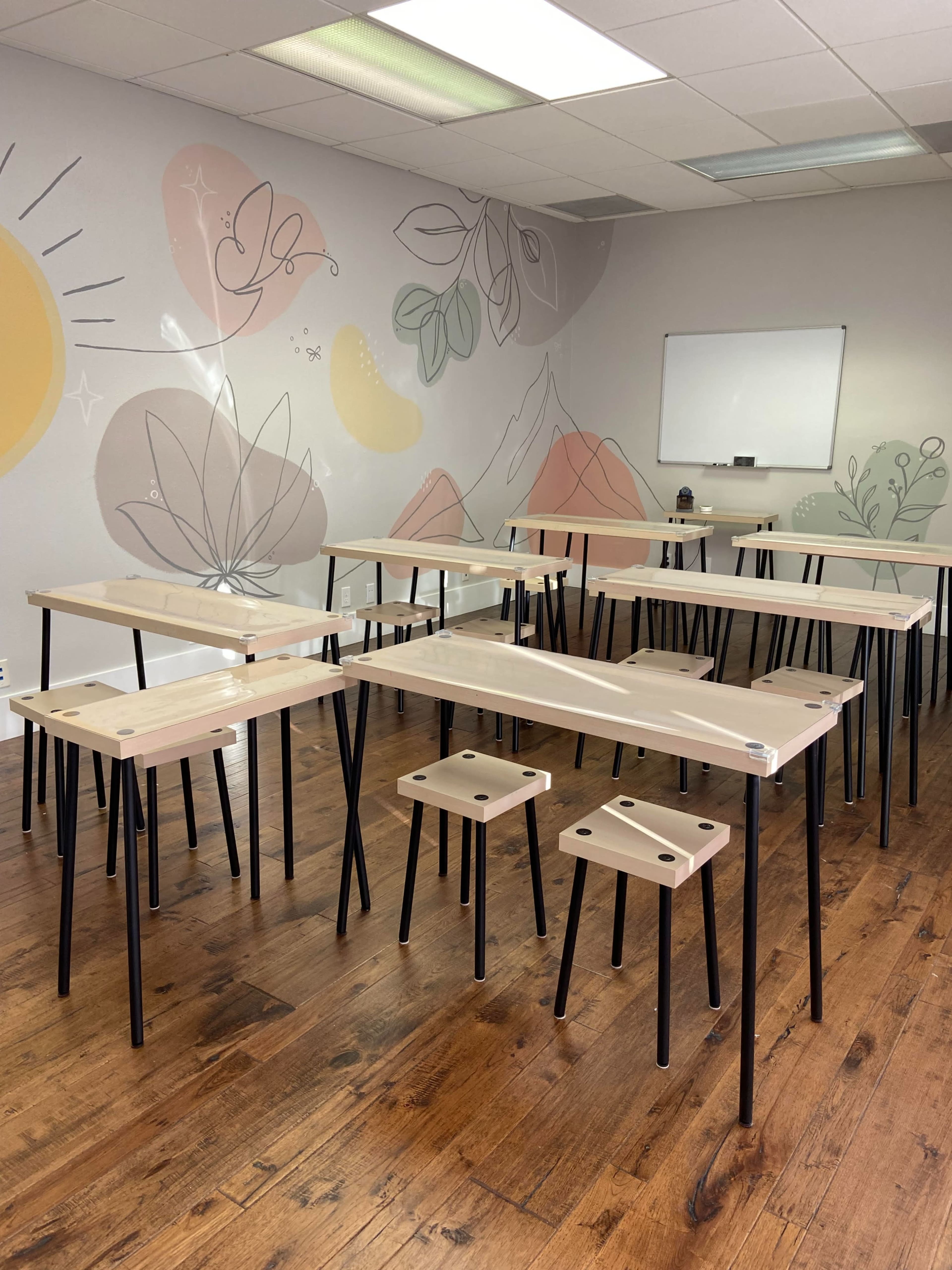 A group of wooden tables with black legs is arranged in a brightly lit room featuring a mural of plants and mountains on the wall.