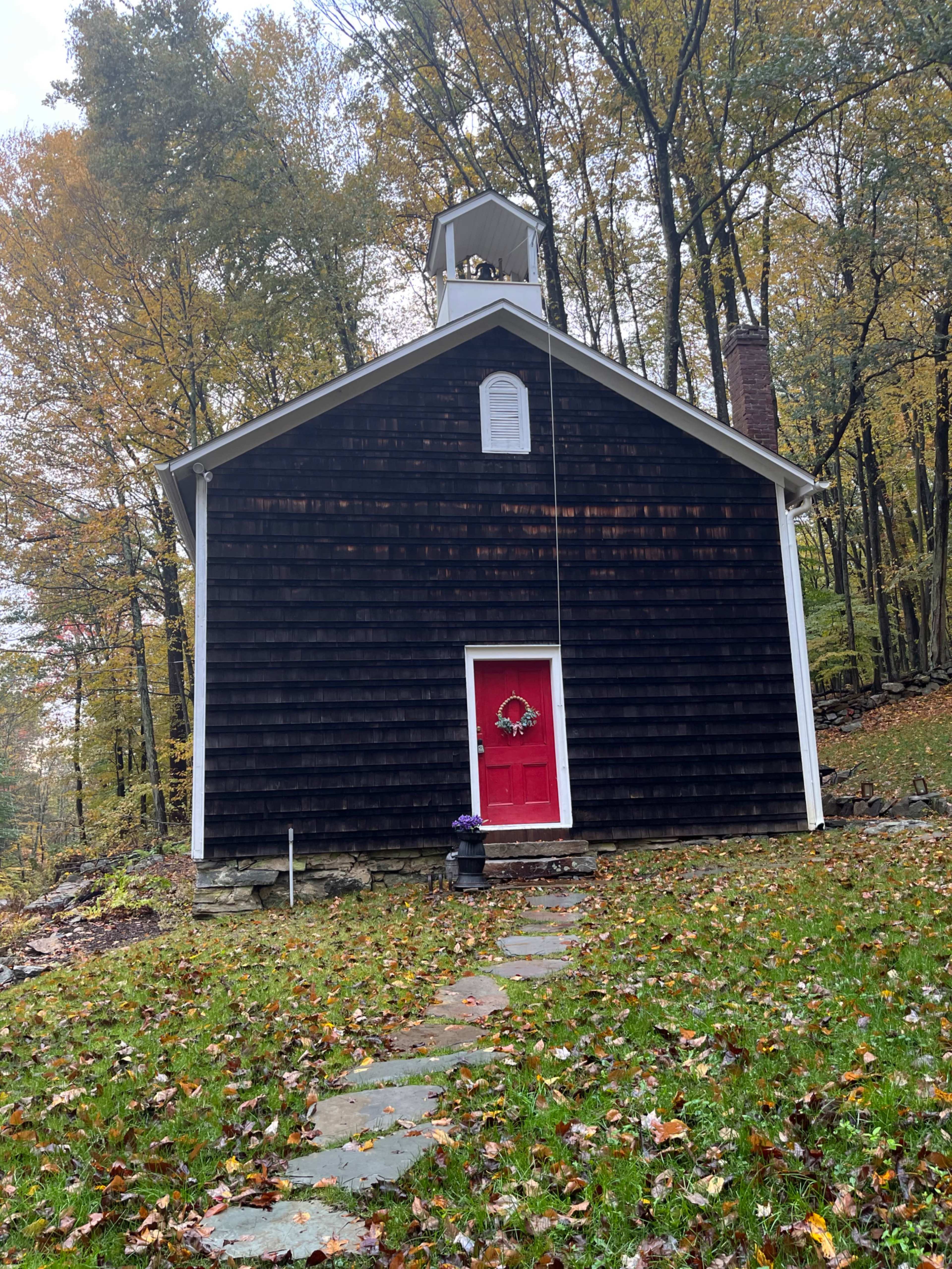 The image shows a dark wood-paneled building with a red door and a bell tower, surrounded by trees and a path lined with stones.