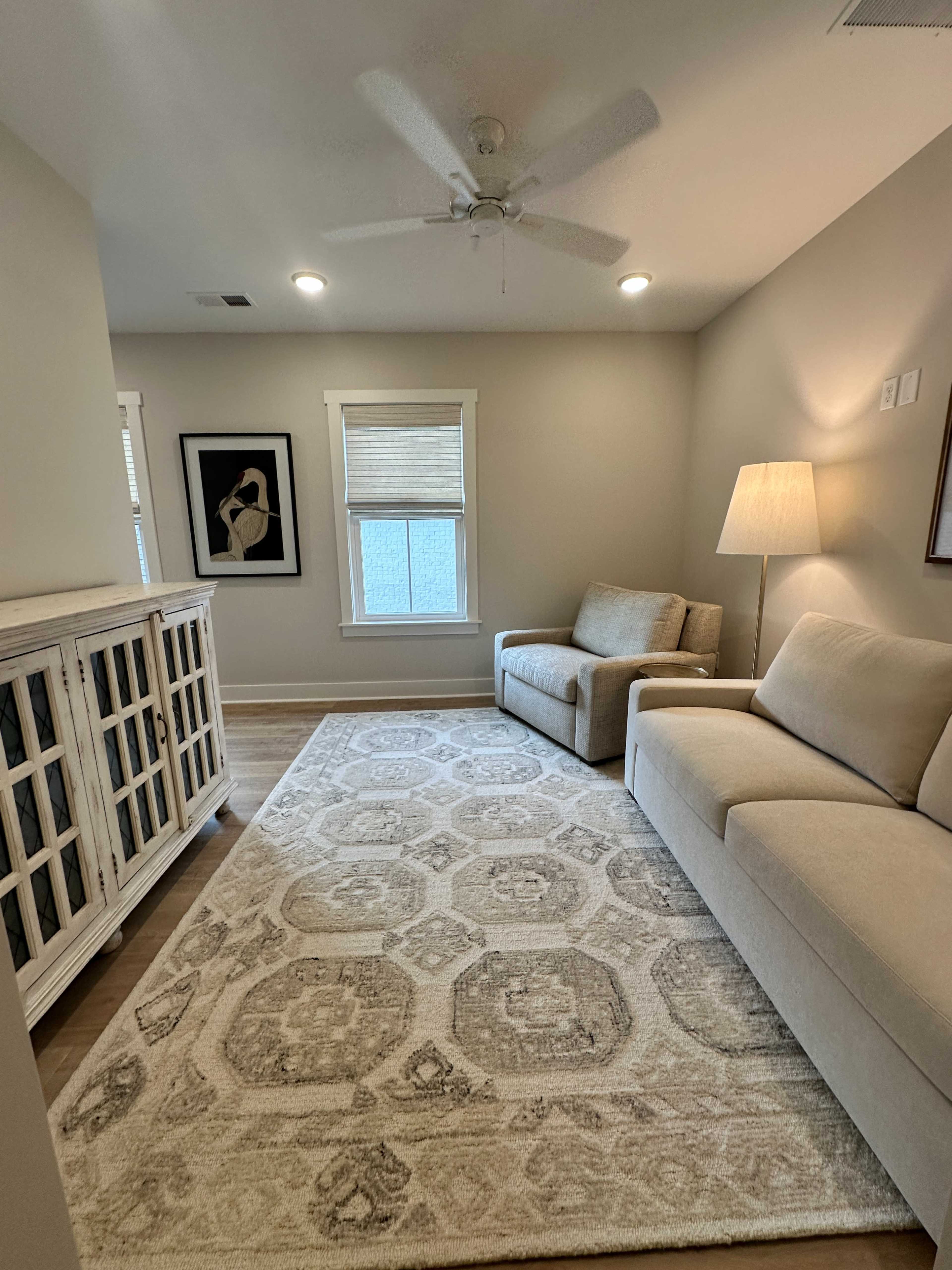The image shows a well-lit living room with a light-colored sofa, armchair, a lamp, and a decorative cabinet on a patterned rug.