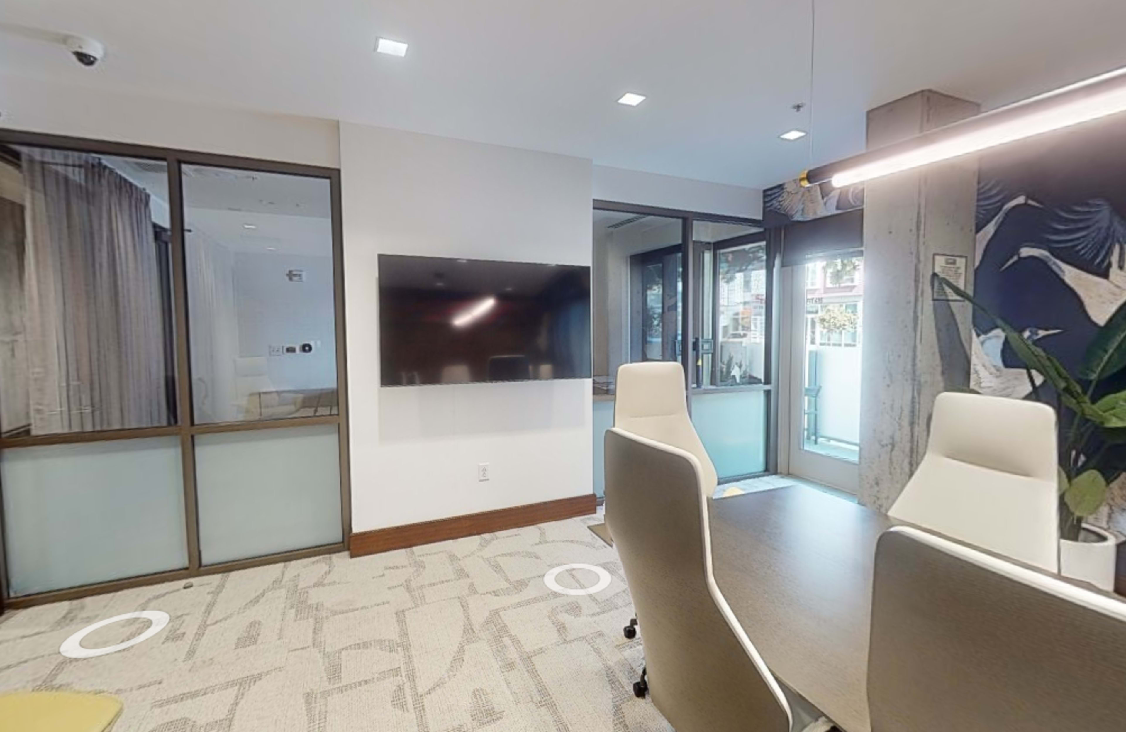 A modern office meeting room featuring a wall-mounted television, two white ergonomic chairs, and large windows allowing natural light.