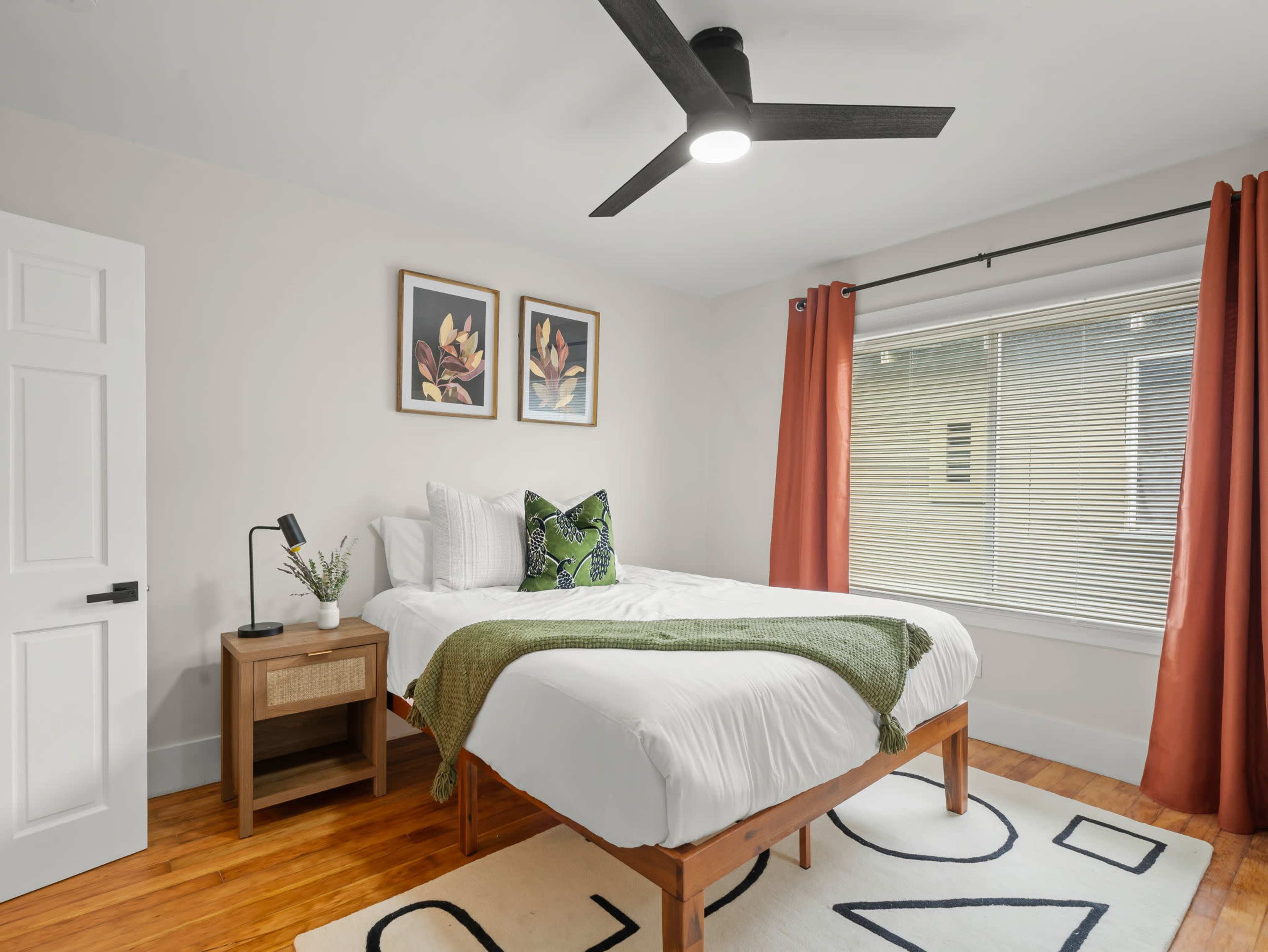 A well-lit bedroom featuring a bed with white linens, green accents, a bedside table, and artwork on the walls.