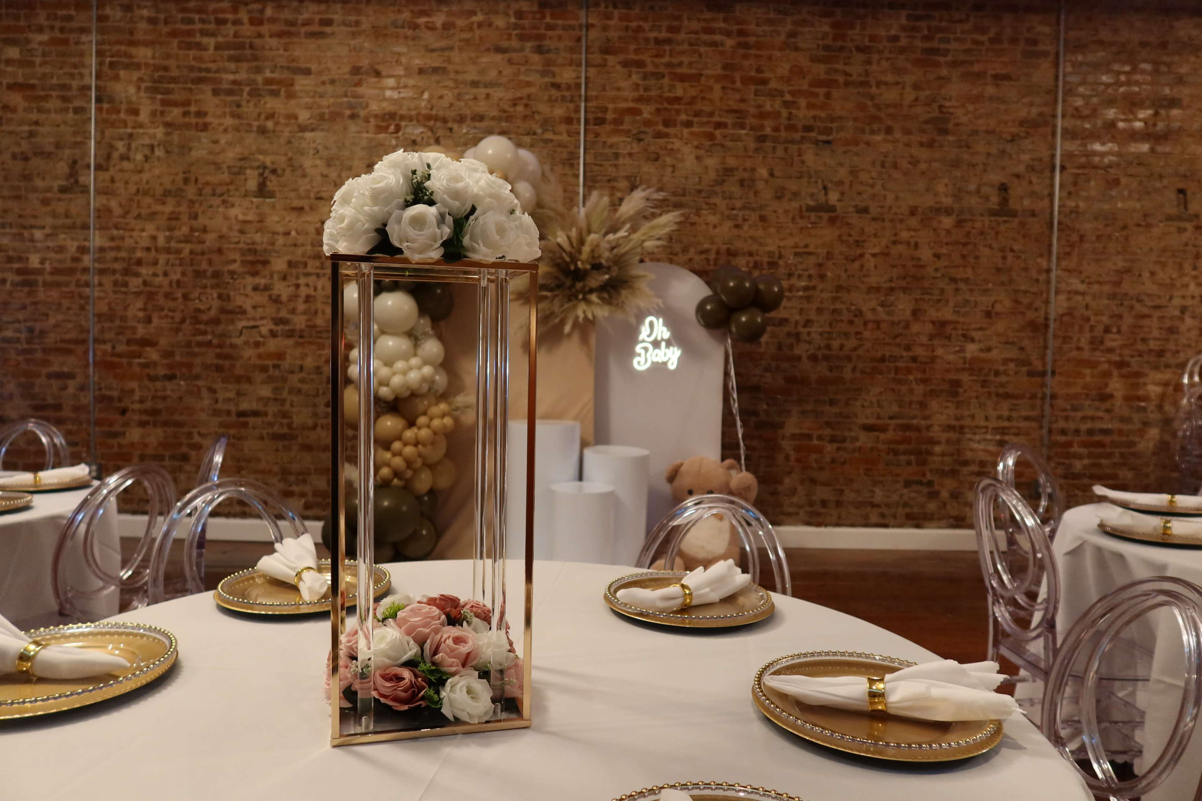 The image shows a decorated event space featuring a centerpiece with flowers in a tall, glass vase on a round table with white cloth and golden accents, against a backdrop of brick walls.