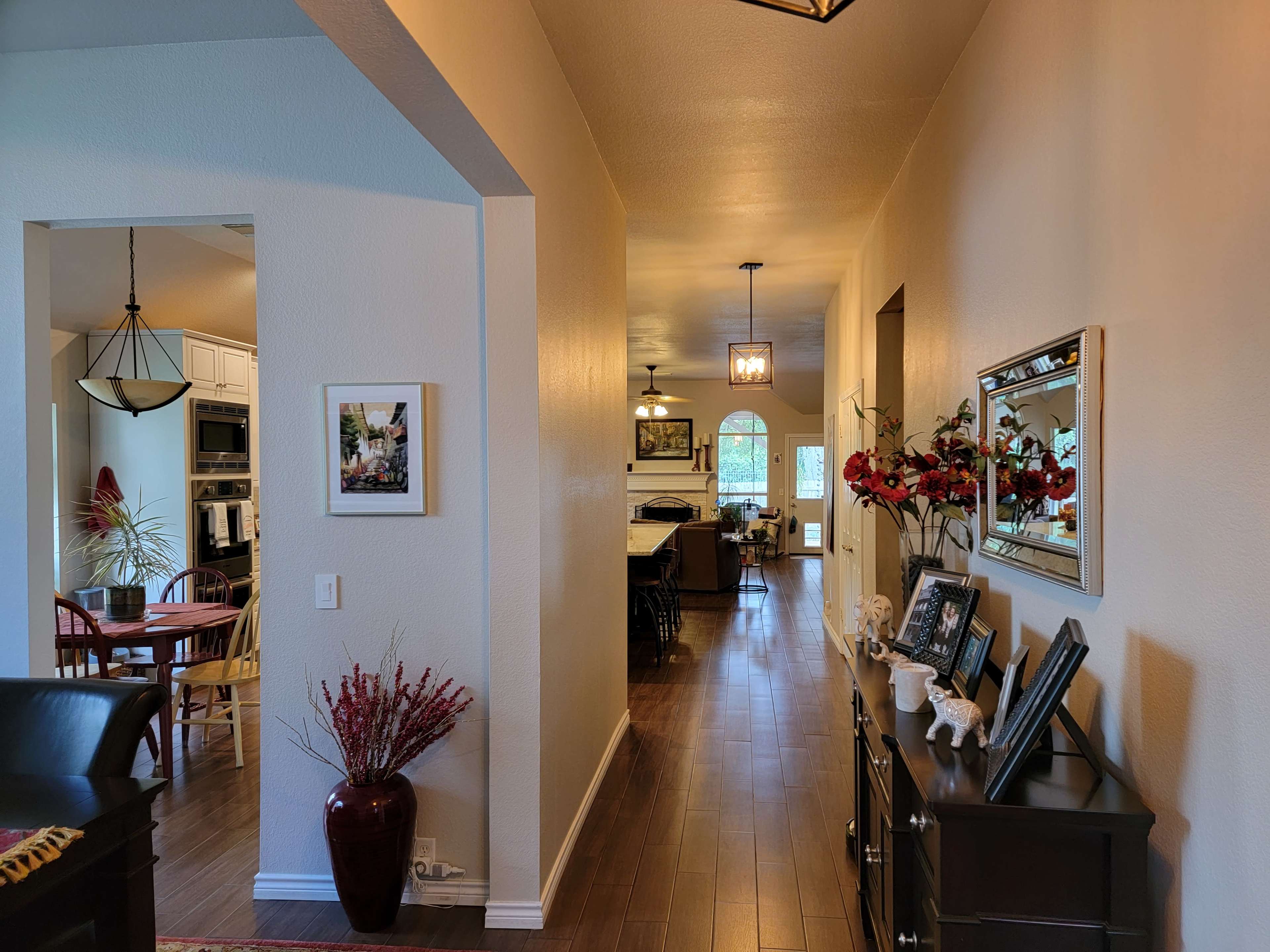 A hallway connects two areas of a home, featuring a table with decorative items and framed photos, while doorways lead to a dining area and a kitchen.
