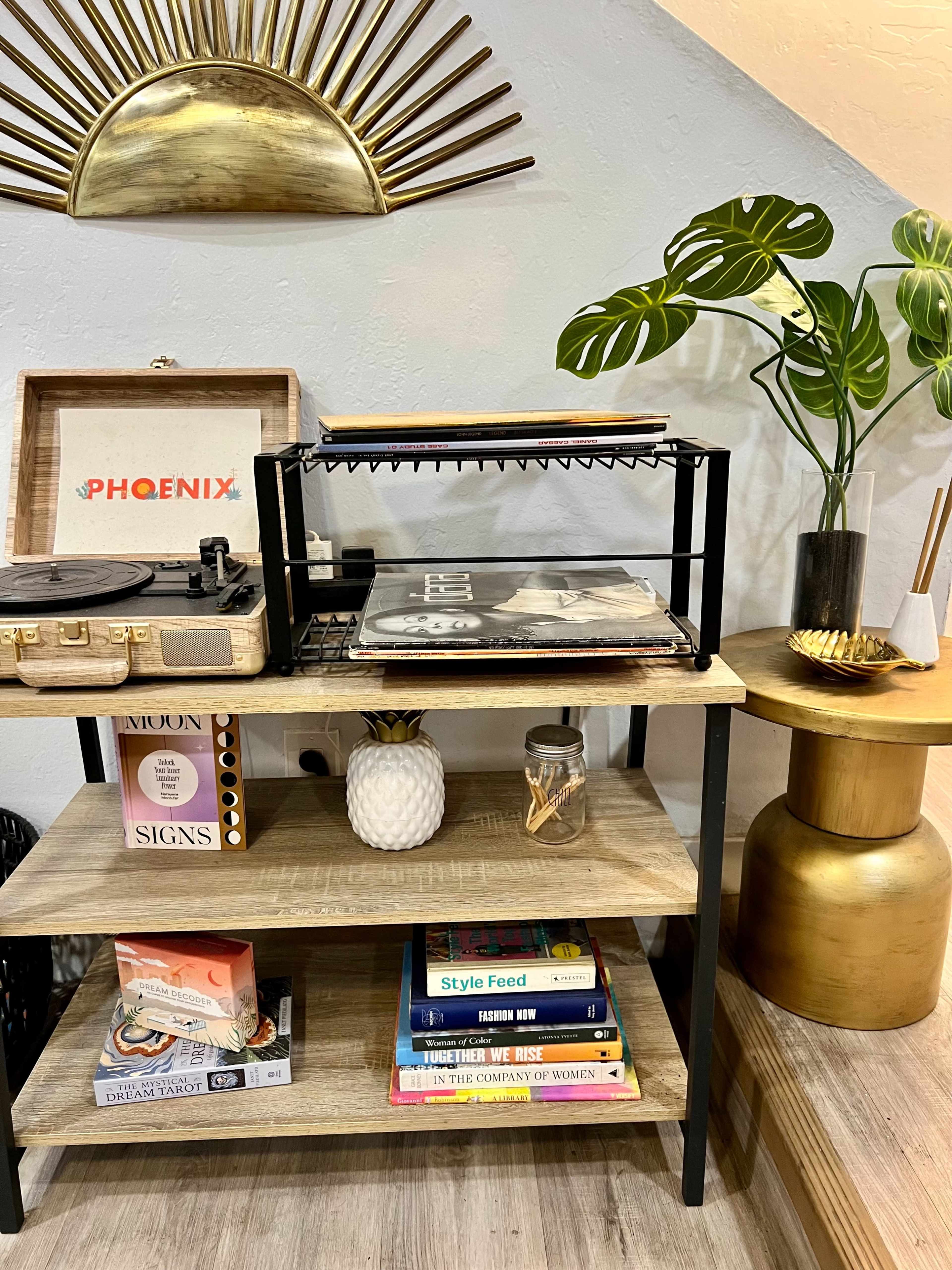 A wooden shelf displays a record player, stacked vinyl records, a pineapple-shaped decoration, and various books beside a tall green plant and a shiny gold table.