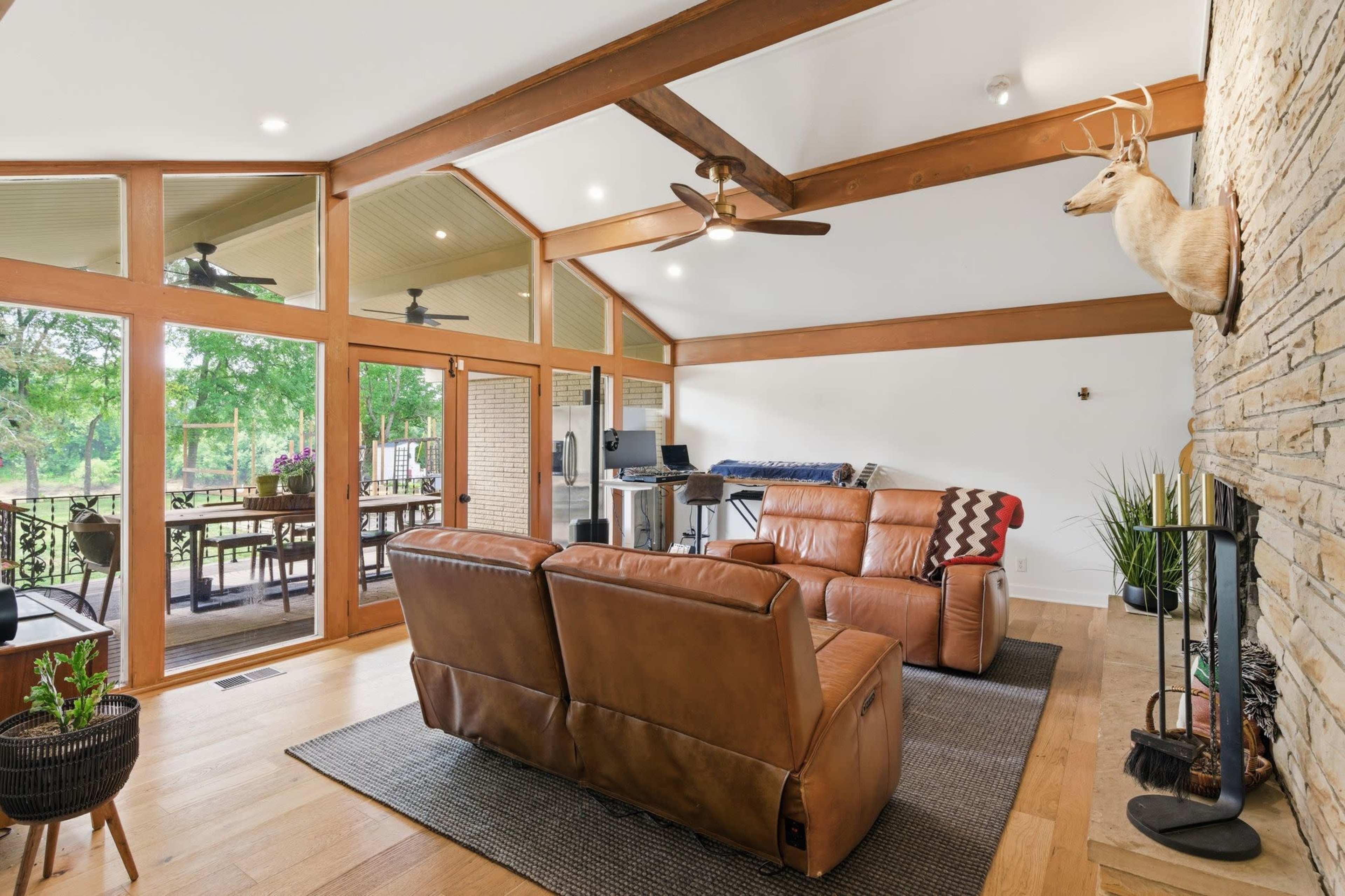 The image shows a modern living room with a leather couch, a stone feature wall, and large windows that open to a deck area surrounded by greenery.