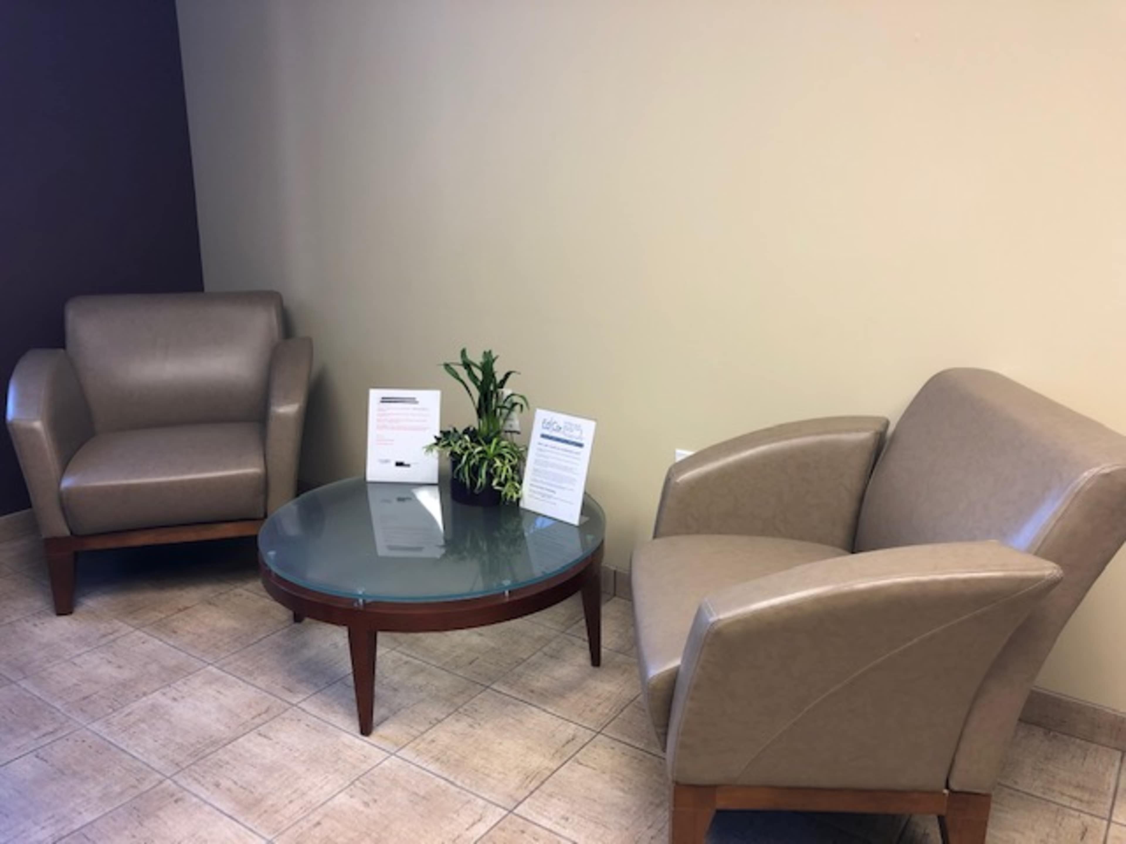 Two beige chairs facing each other around a round glass table, with a small plant on the table and informational pamphlets displayed nearby.