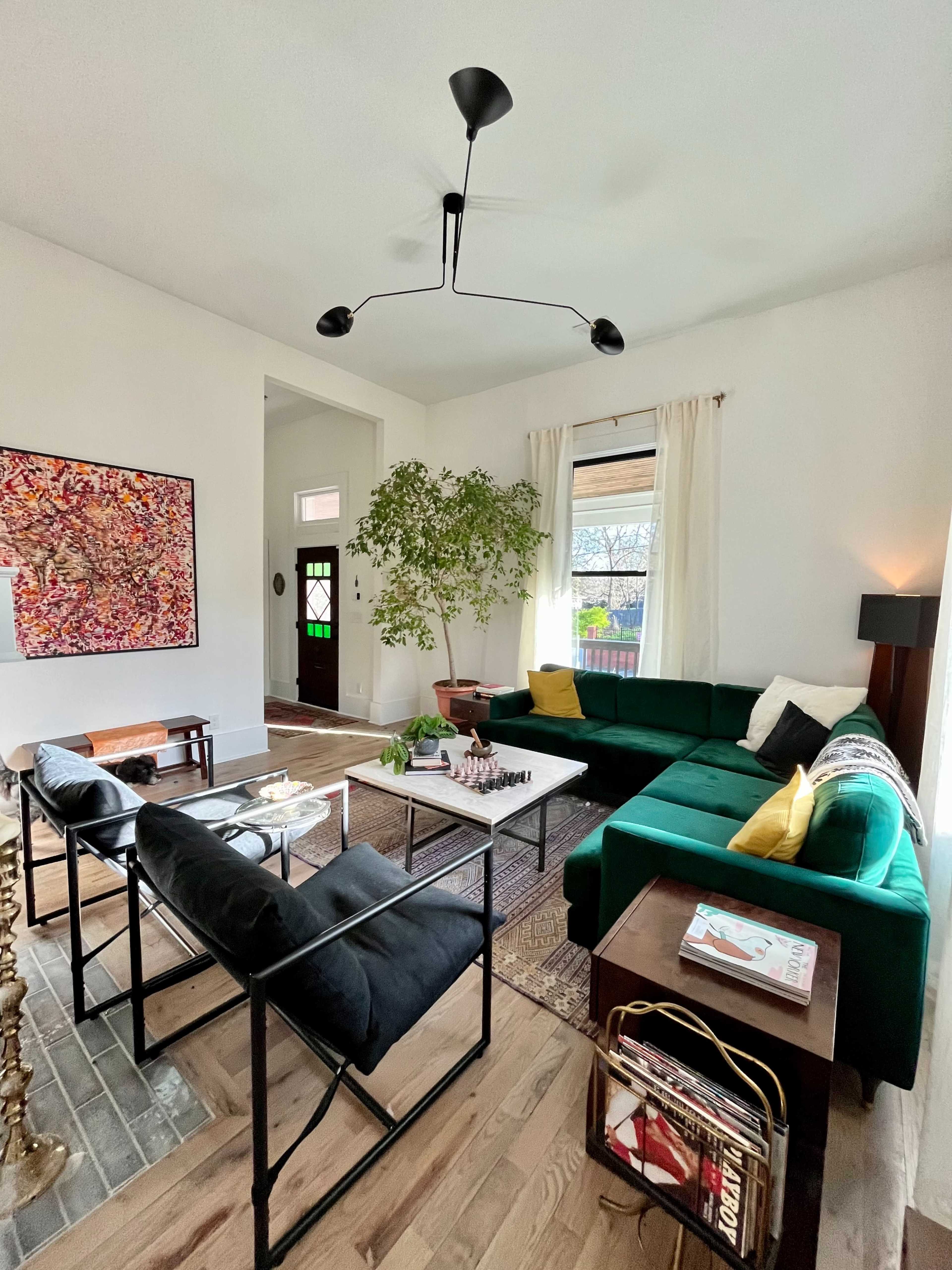 The living room features a green sectional sofa, a coffee table set with a chessboard, and a large window with natural light streaming in.