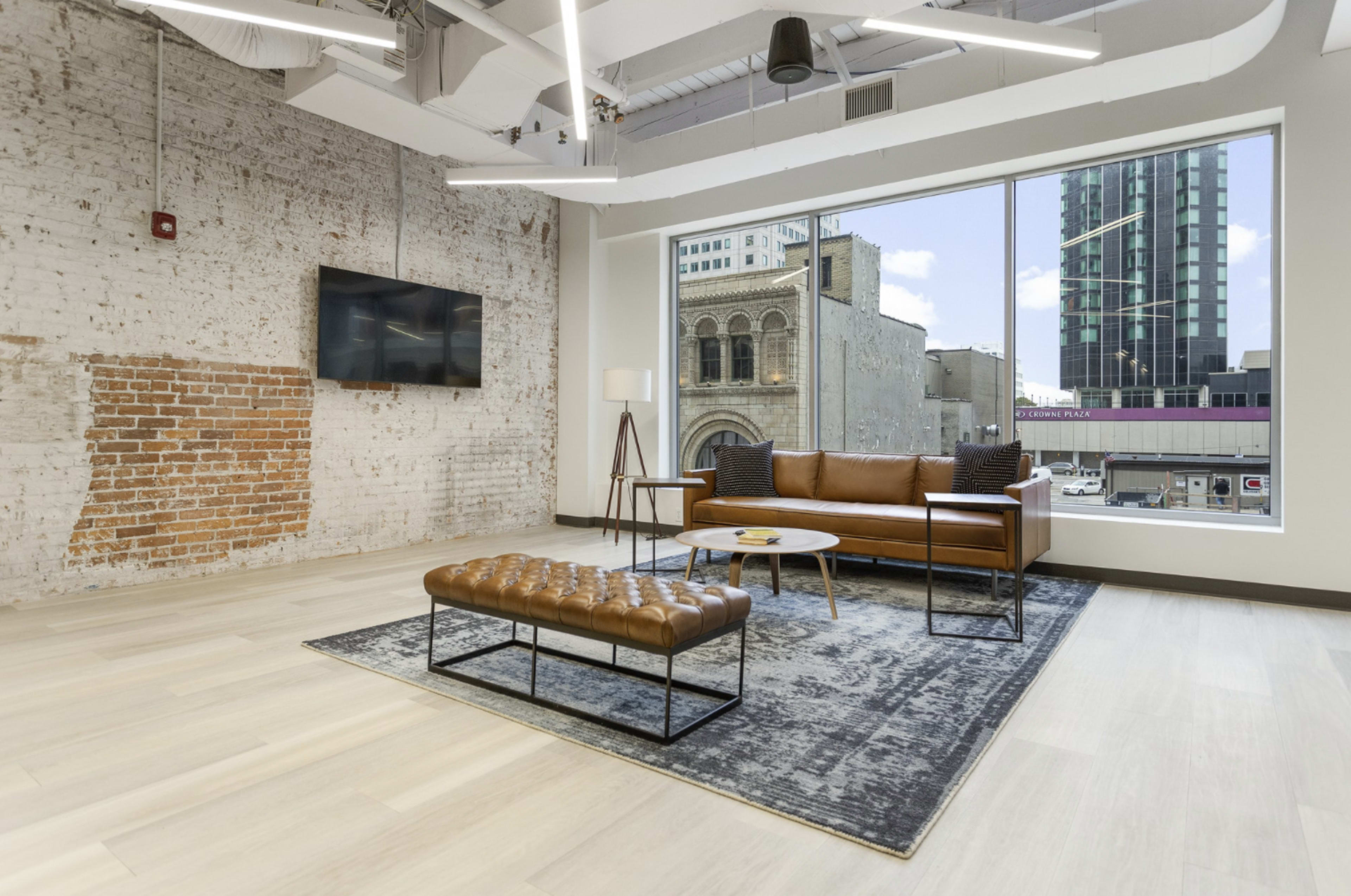 A modern living room with a brown leather sofa, a matching ottoman, a glass coffee table, and large windows overlooking a cityscape, featuring exposed brick walls and wooden flooring.