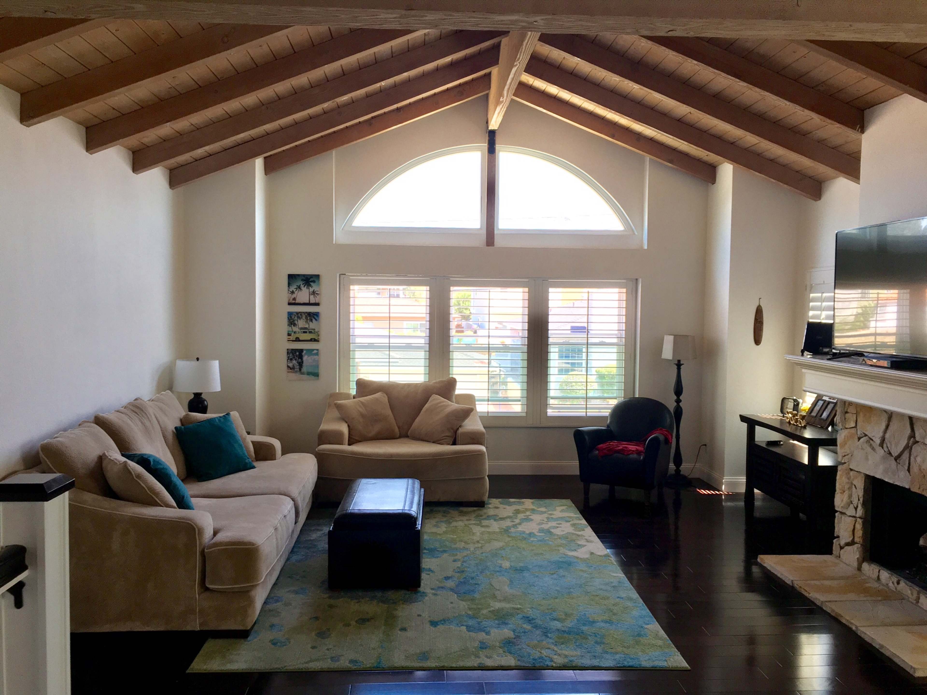 A living room with vaulted wooden ceilings, a beige couch, a light-colored armchair, and a dark coffee table on a colorful rug, all arranged around a stone fireplace and large windows with white shutters.