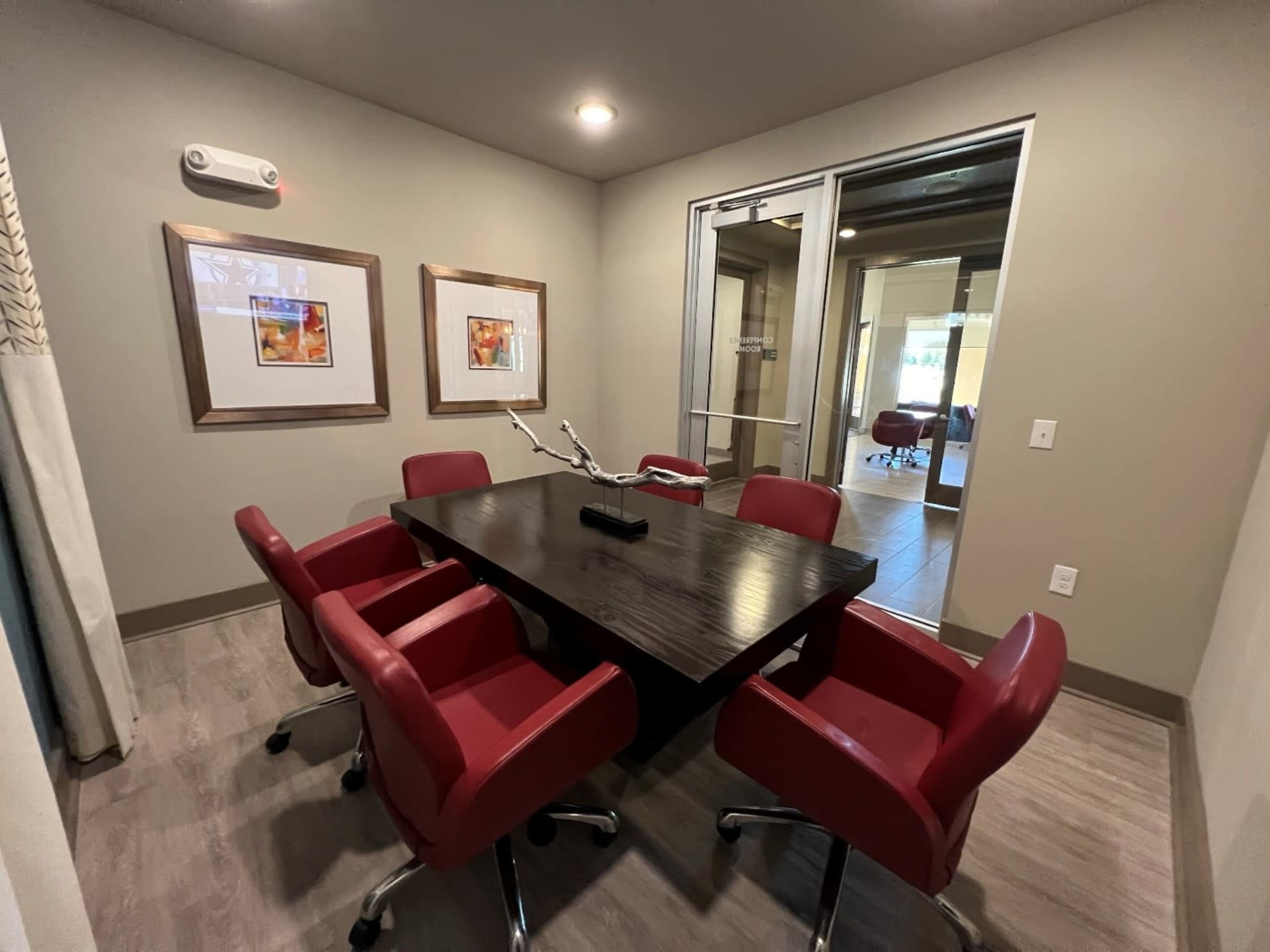 The image depicts a small conference room featuring a rectangular wooden table surrounded by six red chairs, with two framed artworks on the wall and a glass door leading to another space.