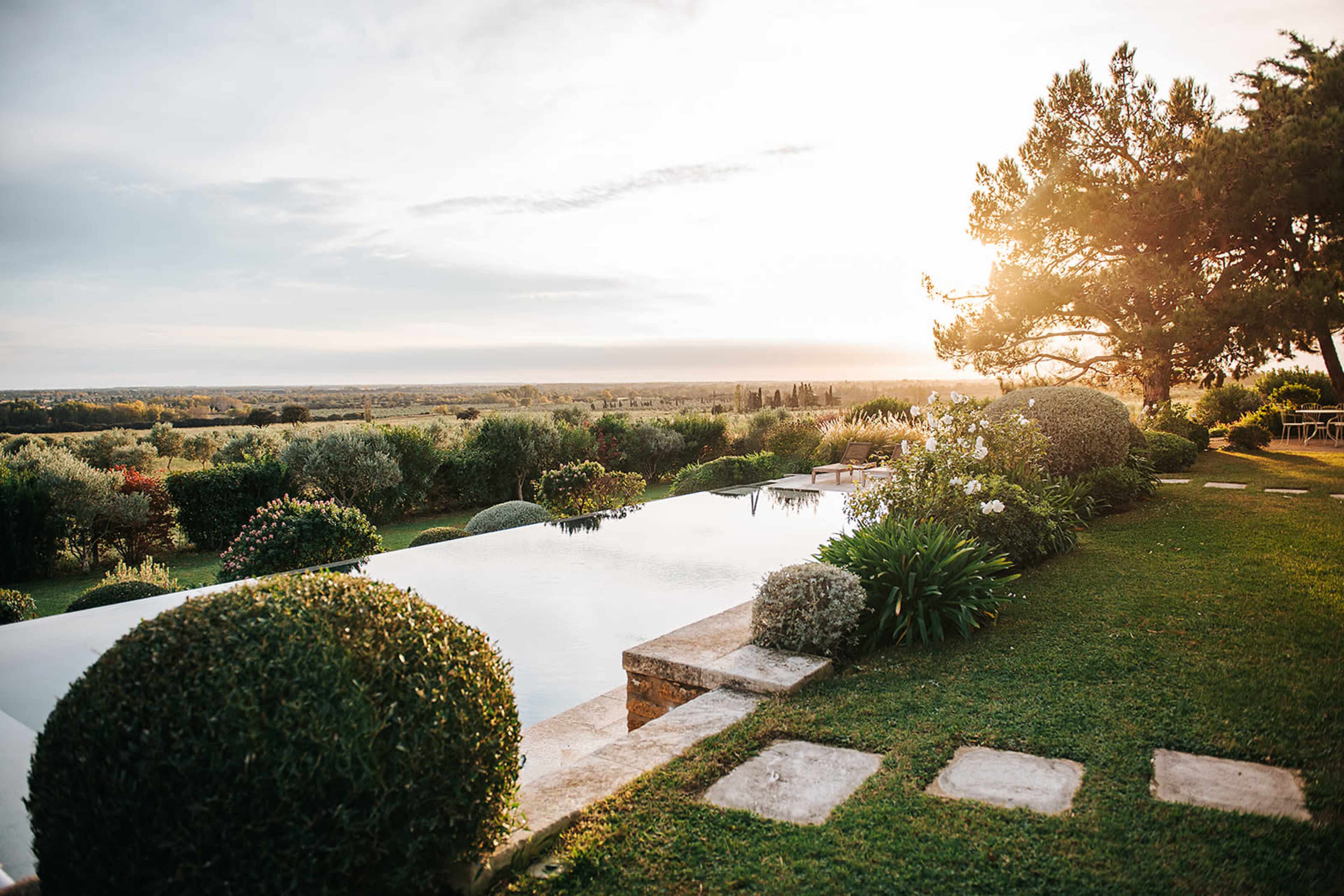 A luxurious infinity pool reflects the sunset, surrounded by neatly trimmed hedges and expansive green fields.
