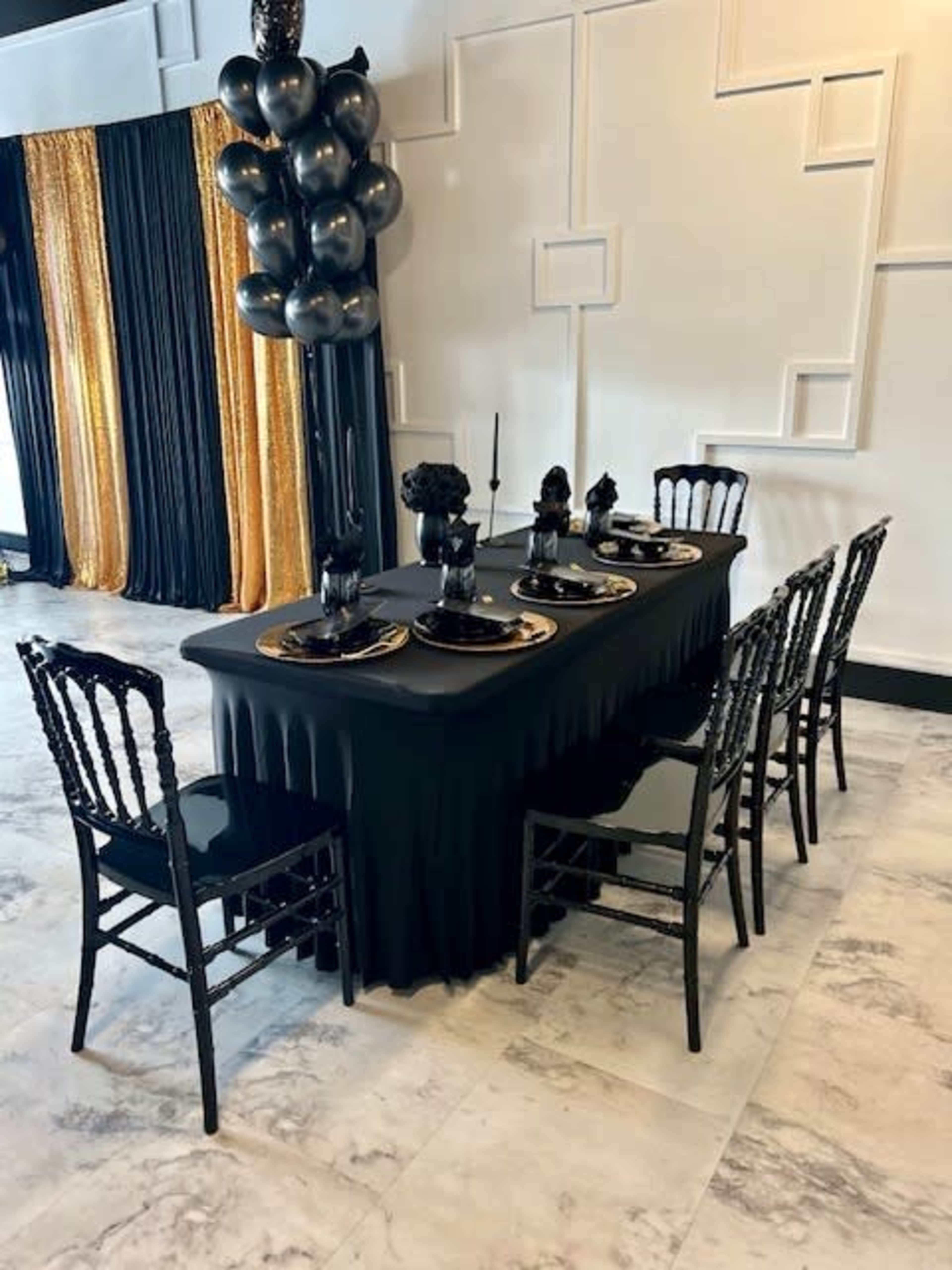 A black table is set with gold-accented plates and black decor, surrounded by matching black chairs in a room with black and gold drapery.