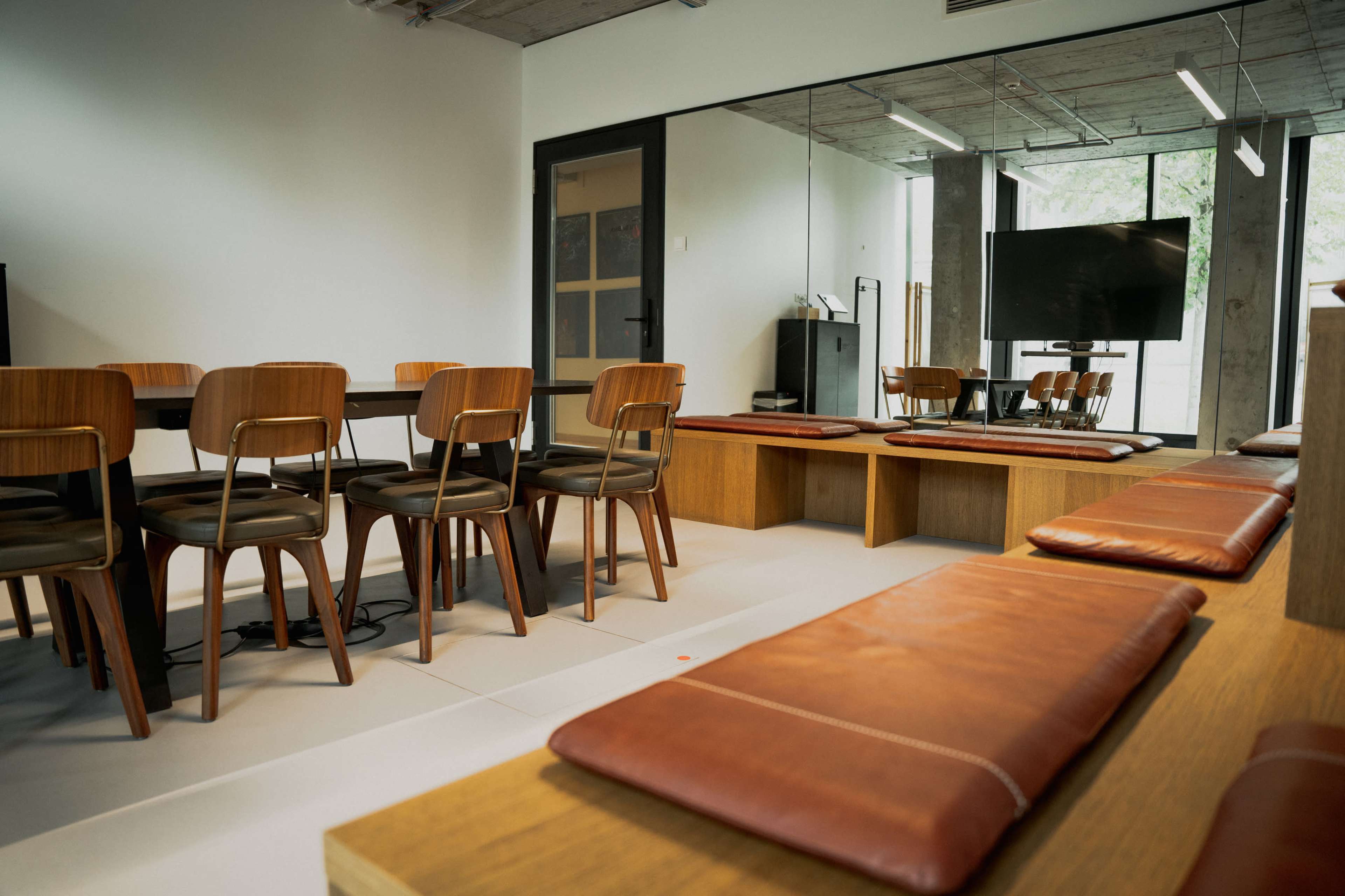 The image shows a modern office space with a seating area featuring wooden chairs and benches, a table, and a large screen in a glass-walled room.