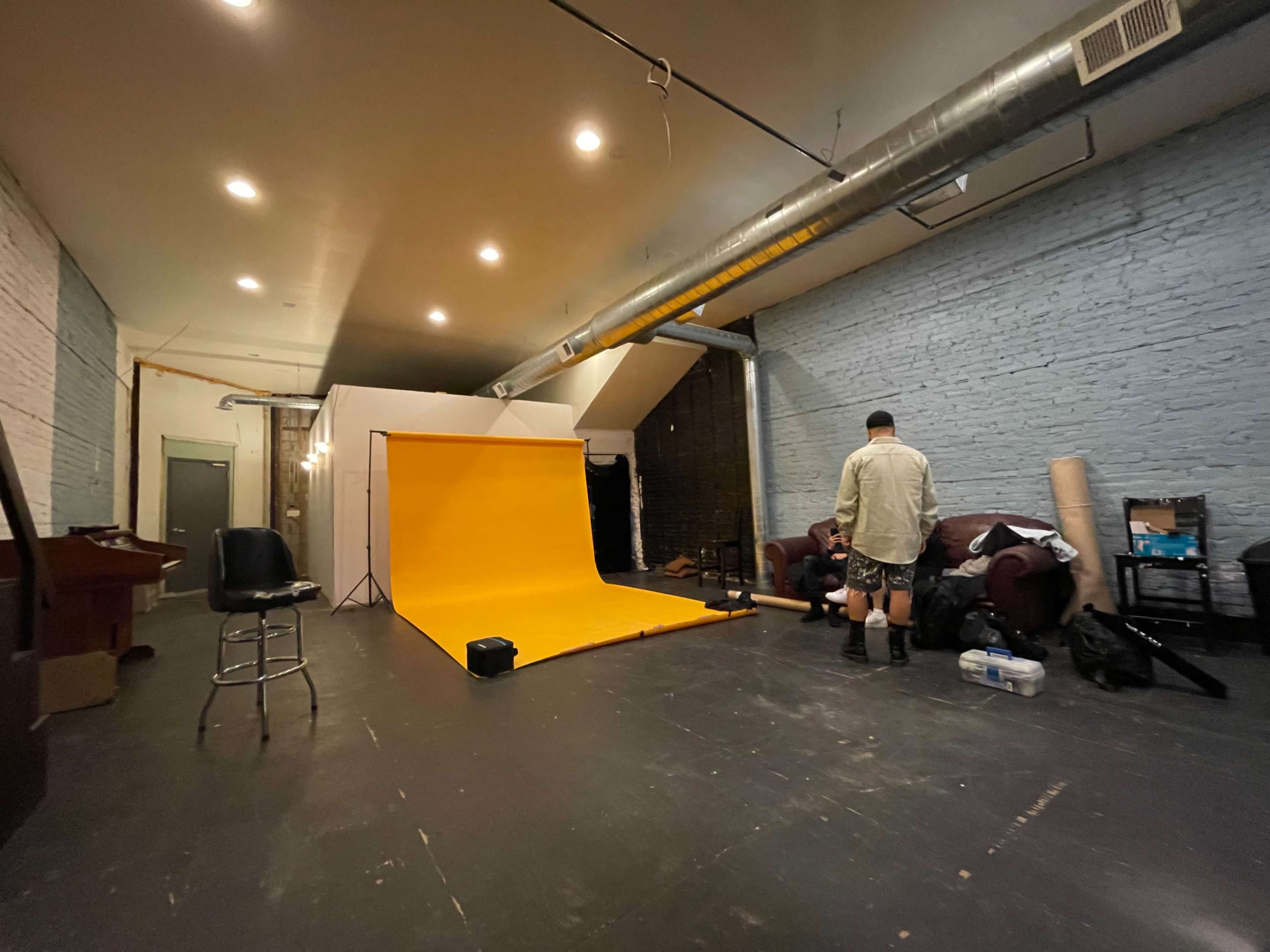 The image shows a spacious studio with a bright orange backdrop, a stool, and several people preparing equipment.