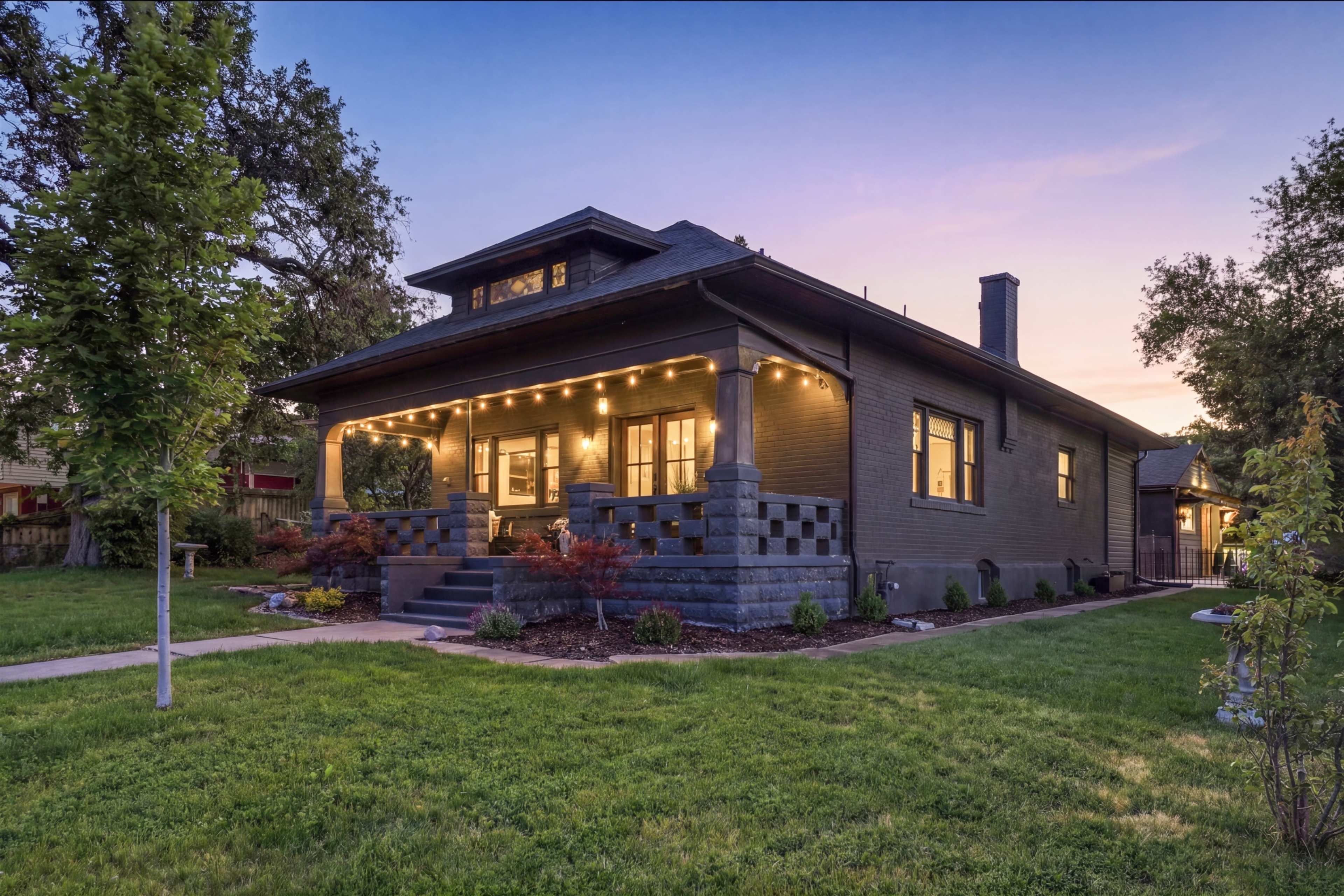 The image shows a two-story house with a landscaped front yard, illuminated by string lights along the porch, at dusk.
