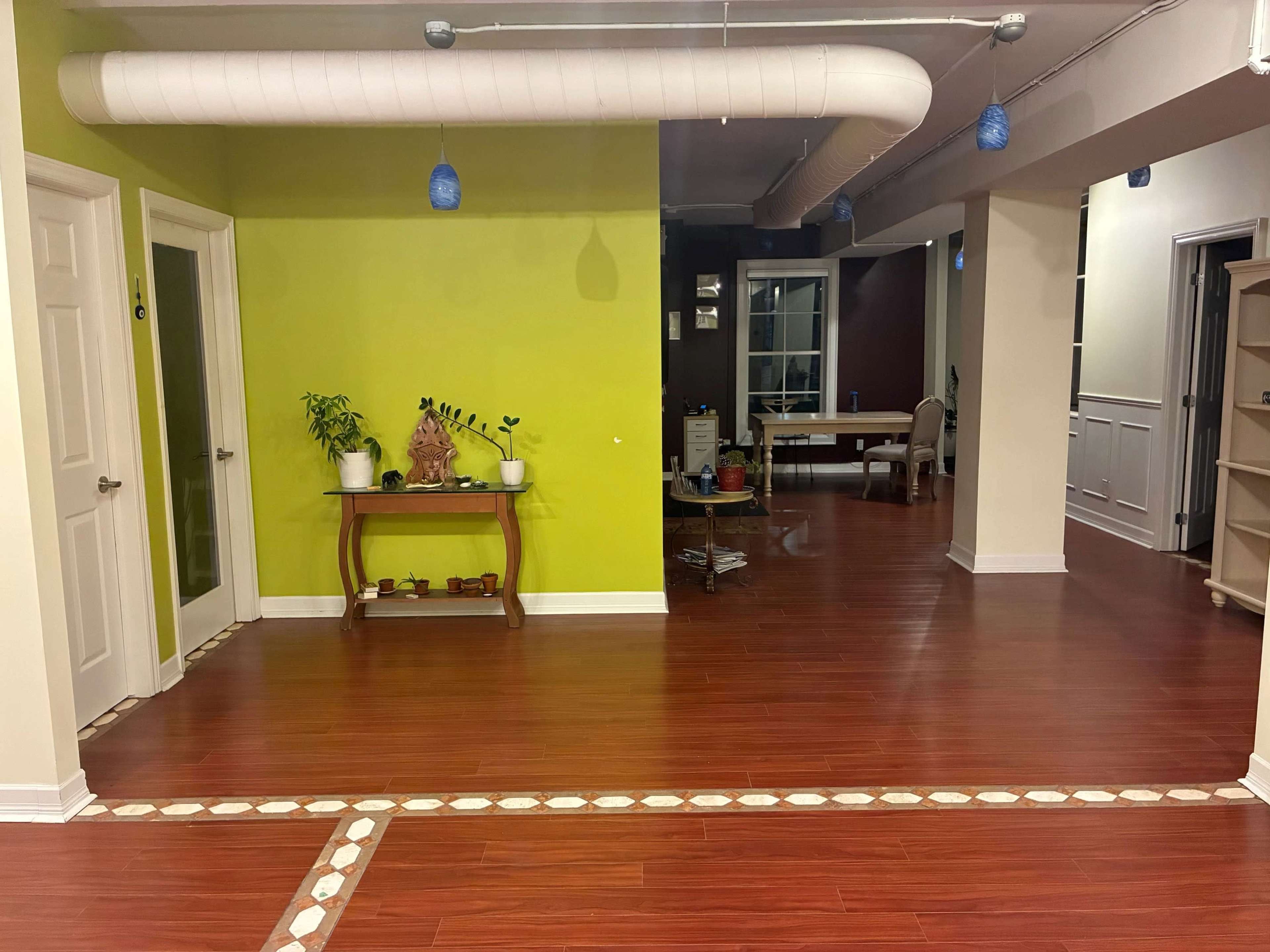 The image shows a spacious room with a green accent wall and wooden flooring, leading to another area with a dining table and various furniture.