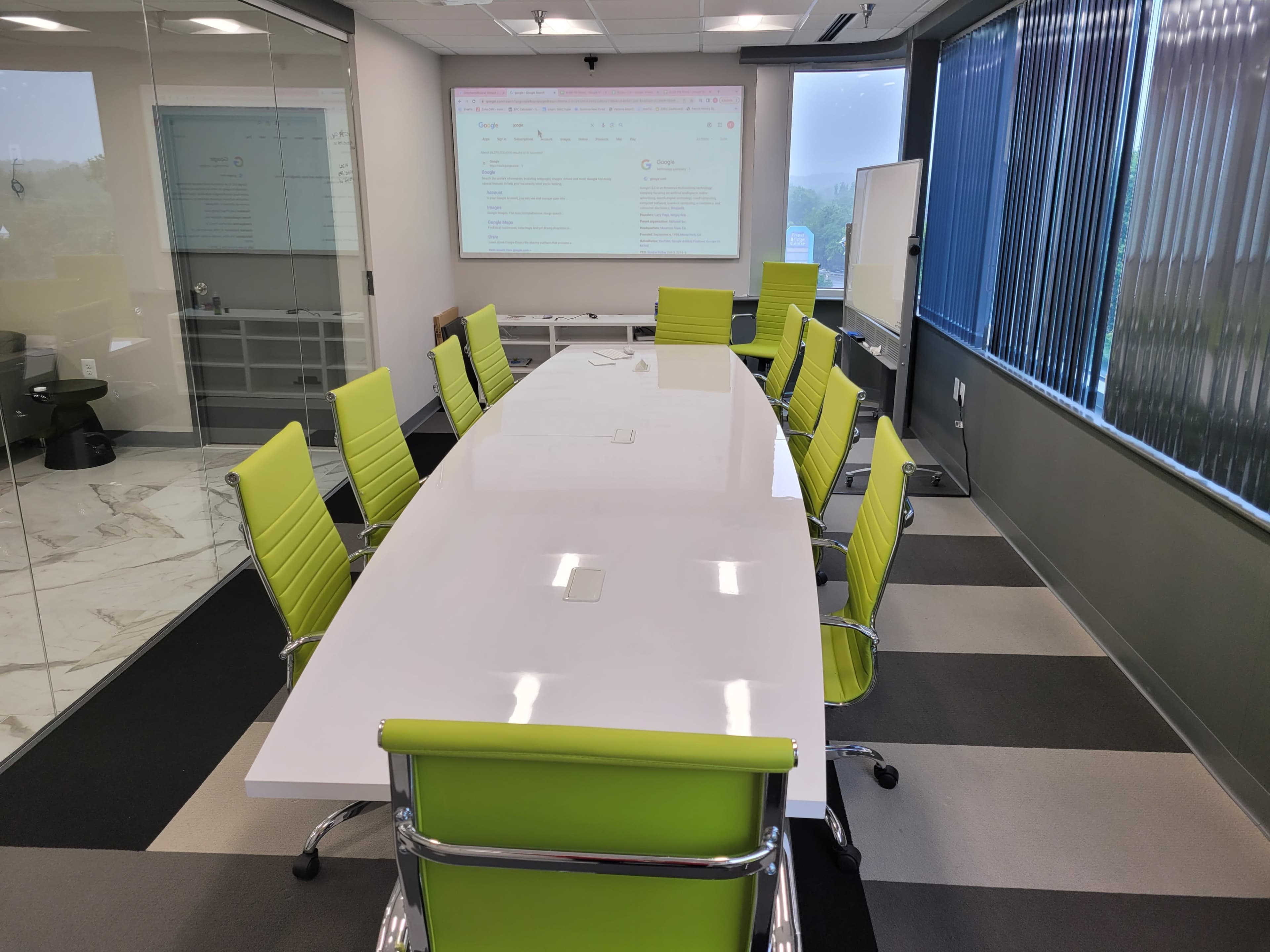 A modern conference room features a long, white table surrounded by green chairs, with a large screen displaying content on one wall.