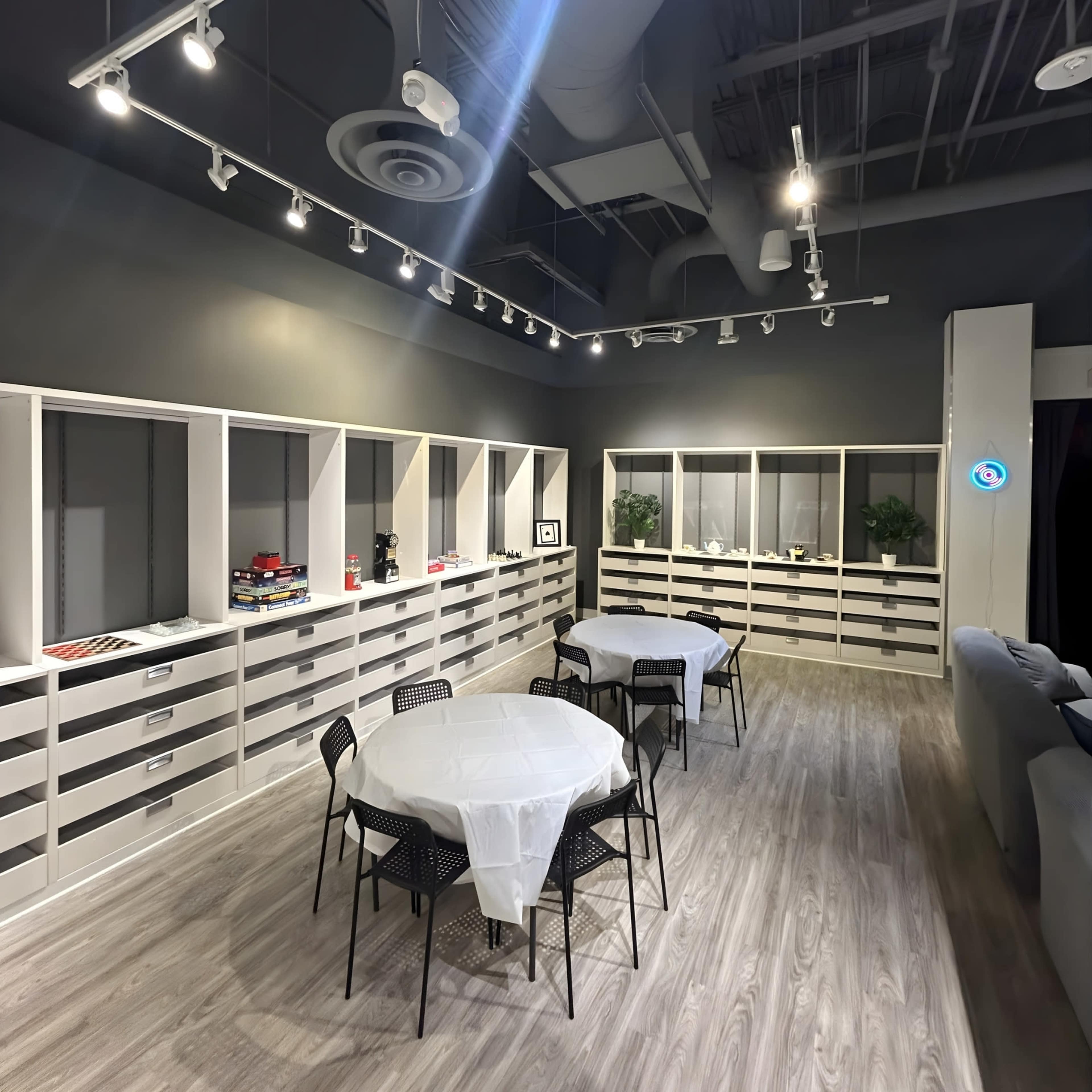 The image shows a spacious, modern room with several white tables and black chairs arranged around them, set against a backdrop of light gray walls and shelving units.