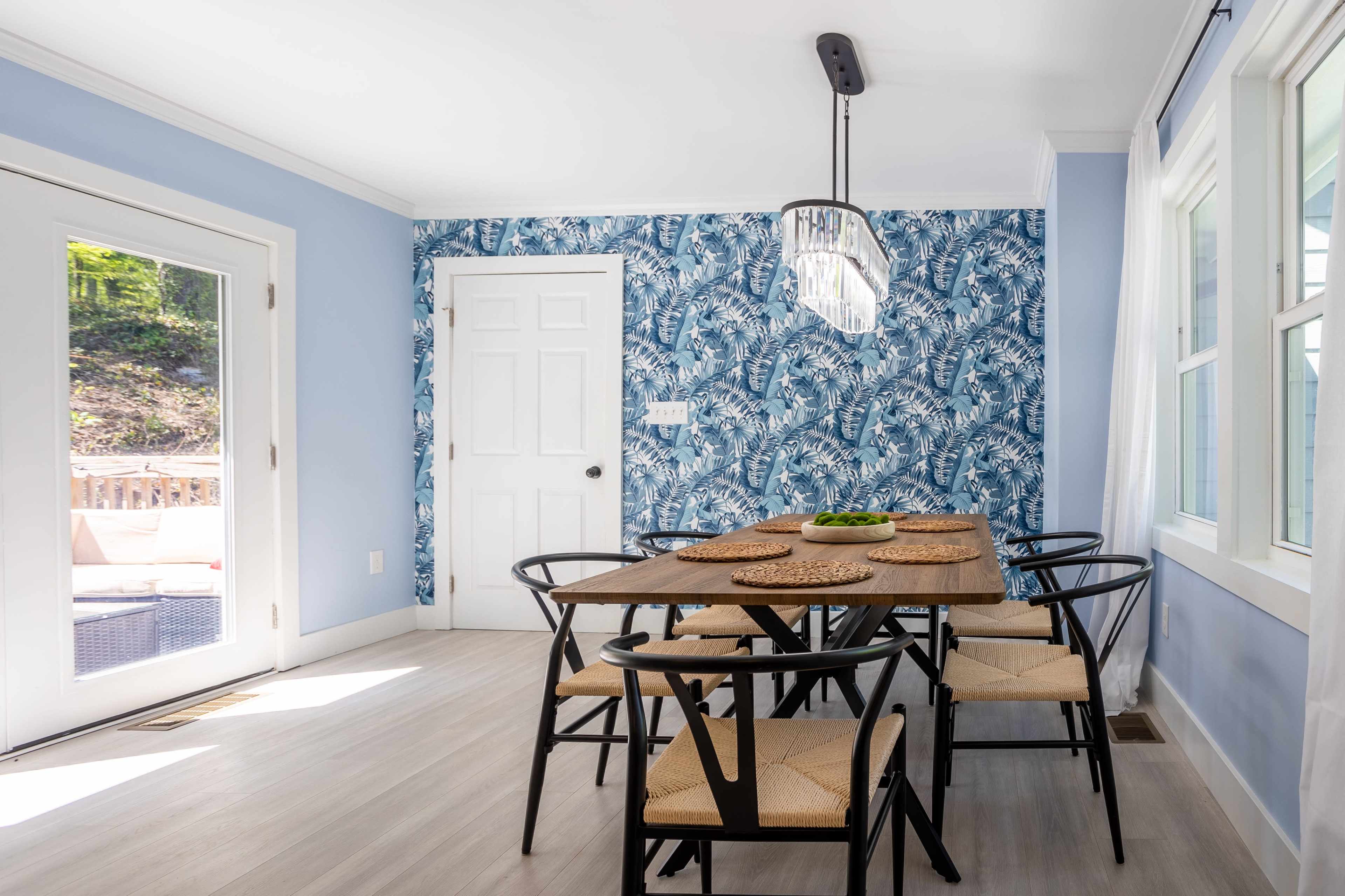 A dining area features a wooden table surrounded by six black chairs, with a blue floral wallpaper and a sliding glass door leading outside.
