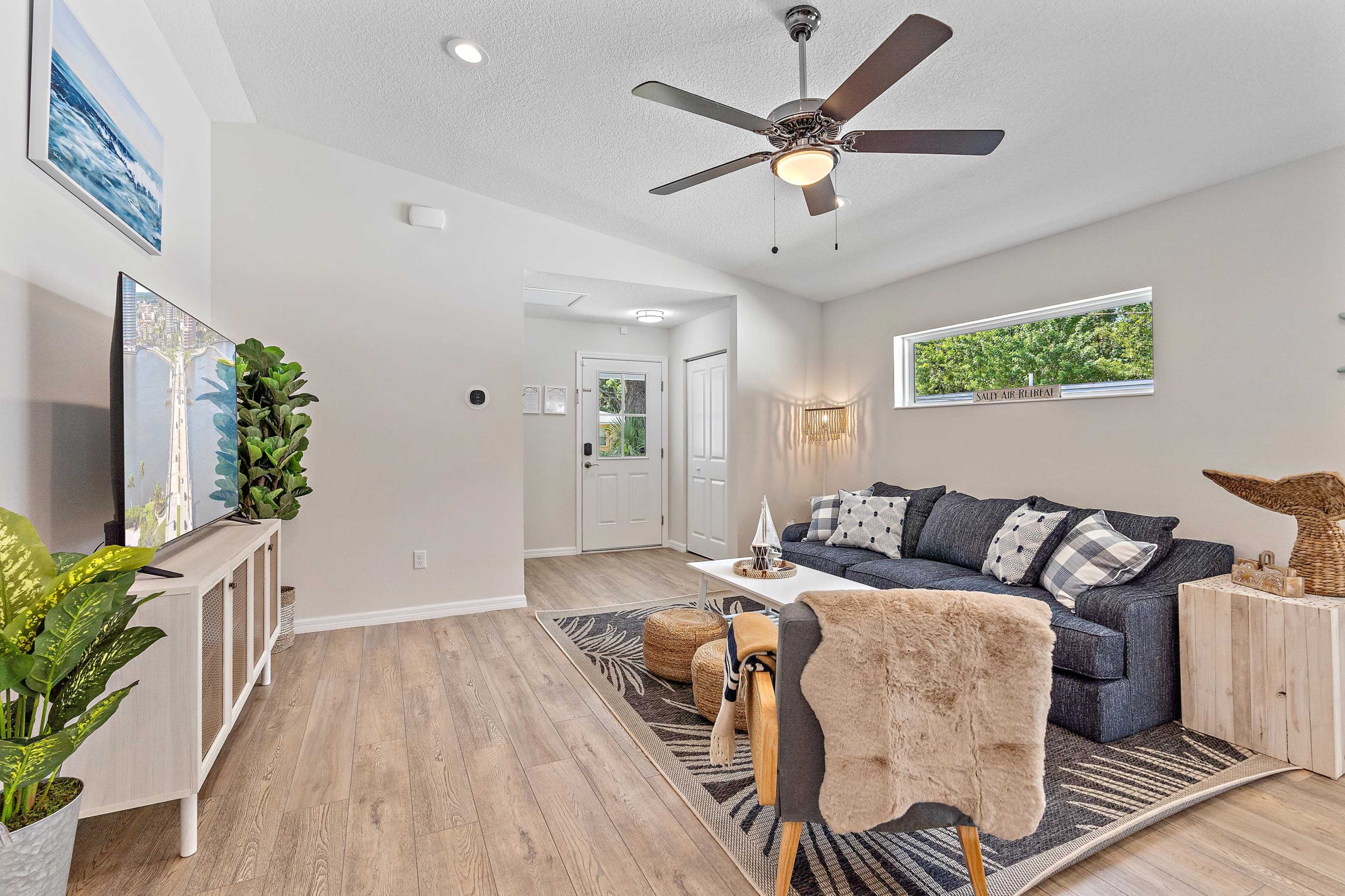 A modern living room features a blue sofa, a wooden coffee table, and a large wall-mounted television, with natural light streaming through a window.