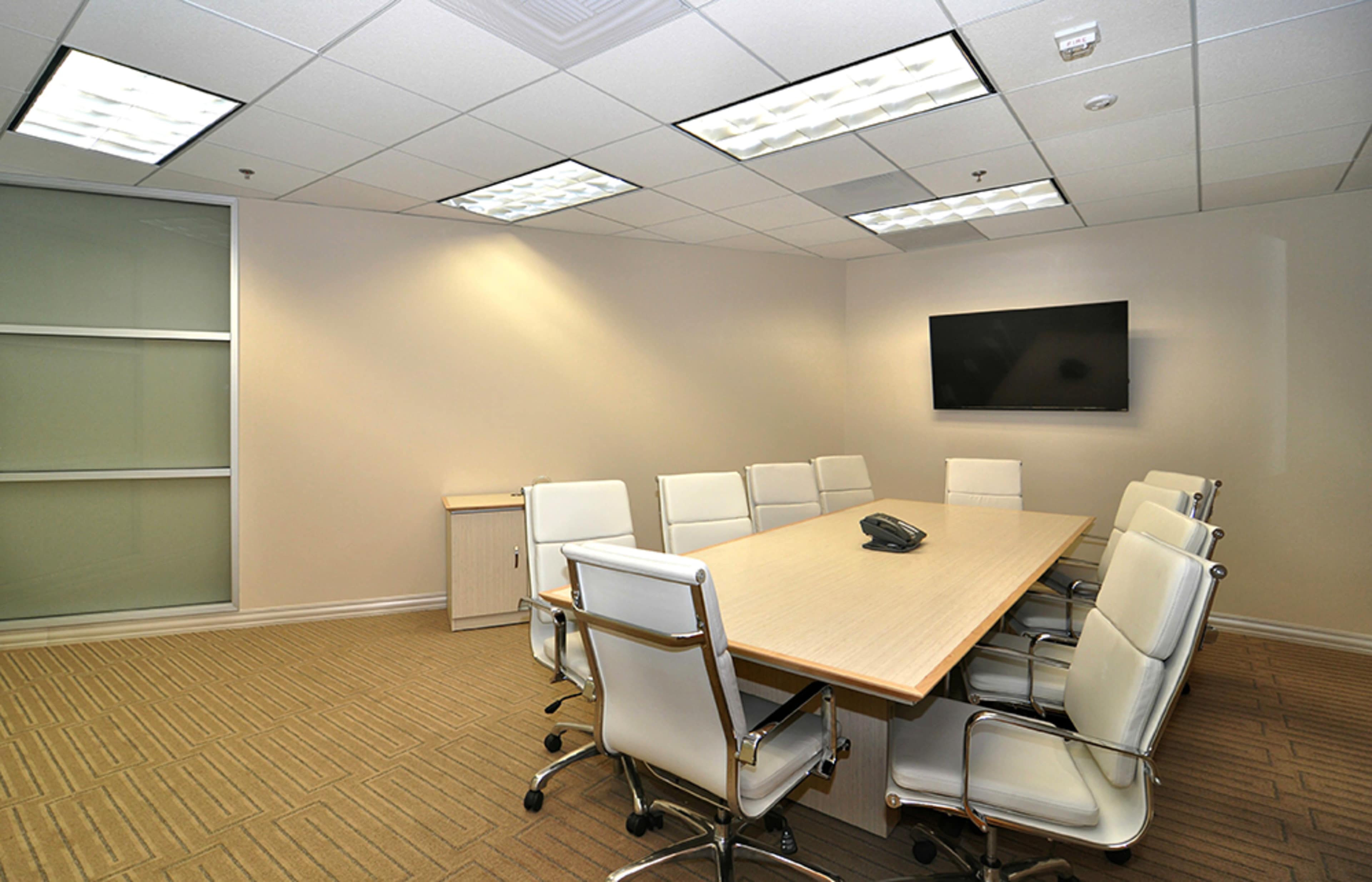 A spacious conference room with a long wooden table surrounded by white chairs, a wall-mounted TV, and large windows.