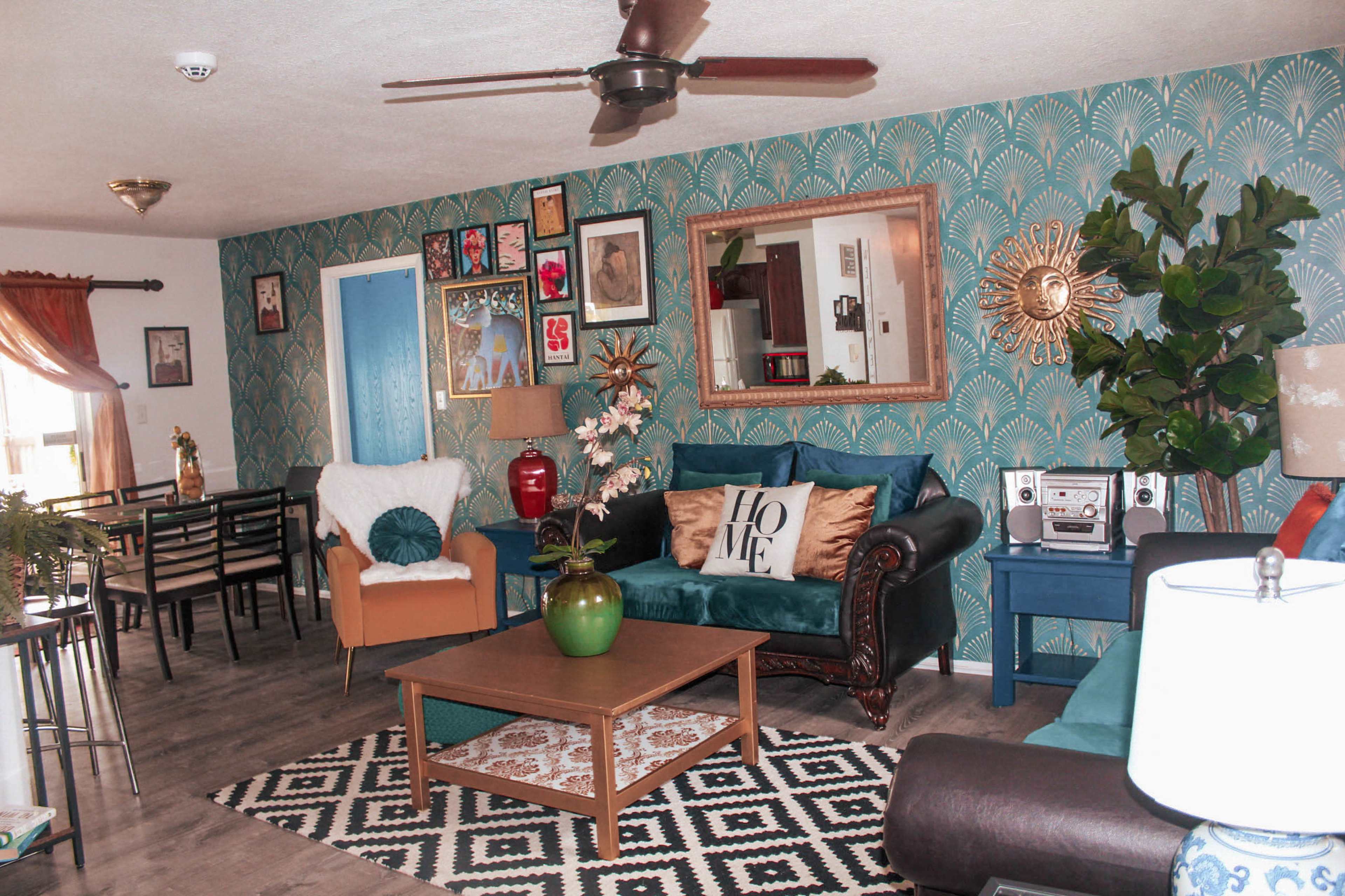 A living room with a patterned teal wallpaper, a mix of furniture including a green sofa and a brown couch, and decorative elements like a coffee table and artwork on the walls.