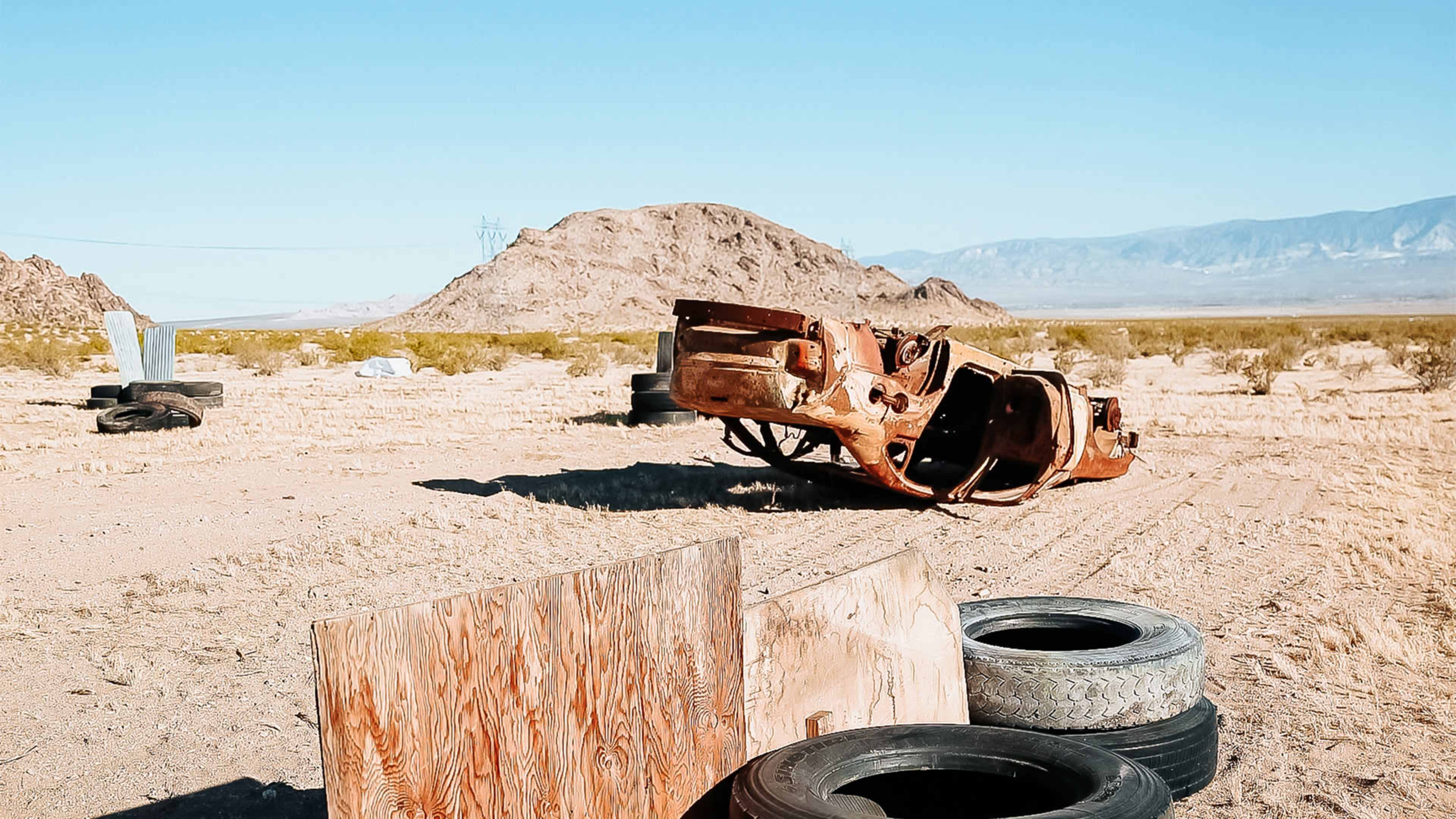 An abandoned, rusted car is overturned in a barren desert landscape, surrounded by wooden planks and old tires.