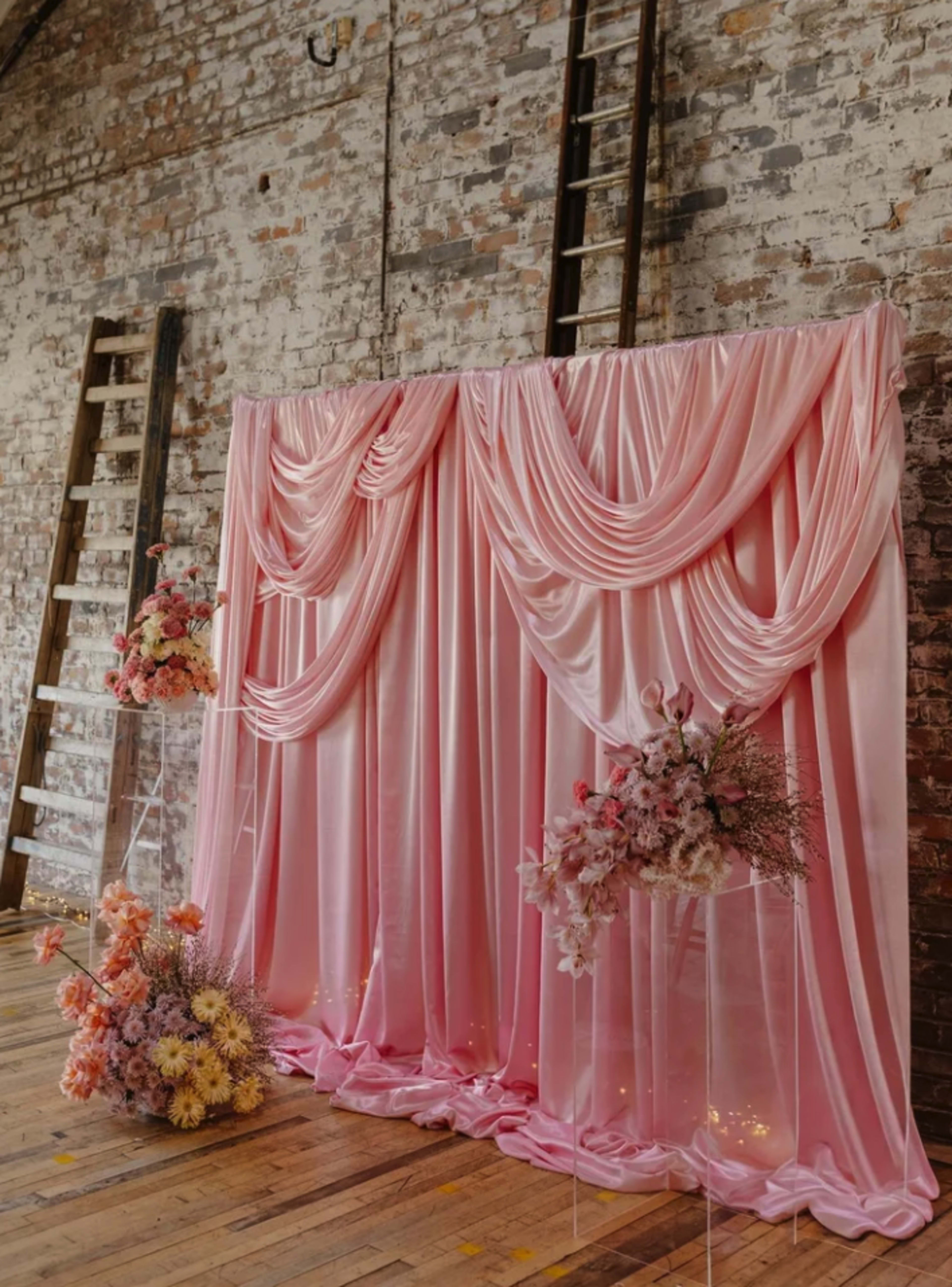 The image features a pink satin backdrop draped elegantly with floral arrangements on clear stands in front of a brick wall.